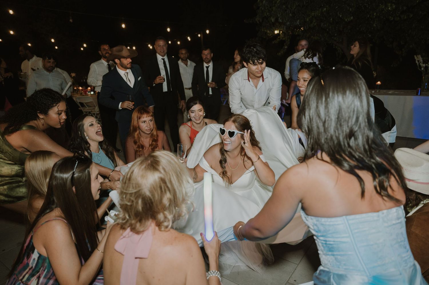 Bride in white dress dancing with guests at lively outdoor wedding reception at night.