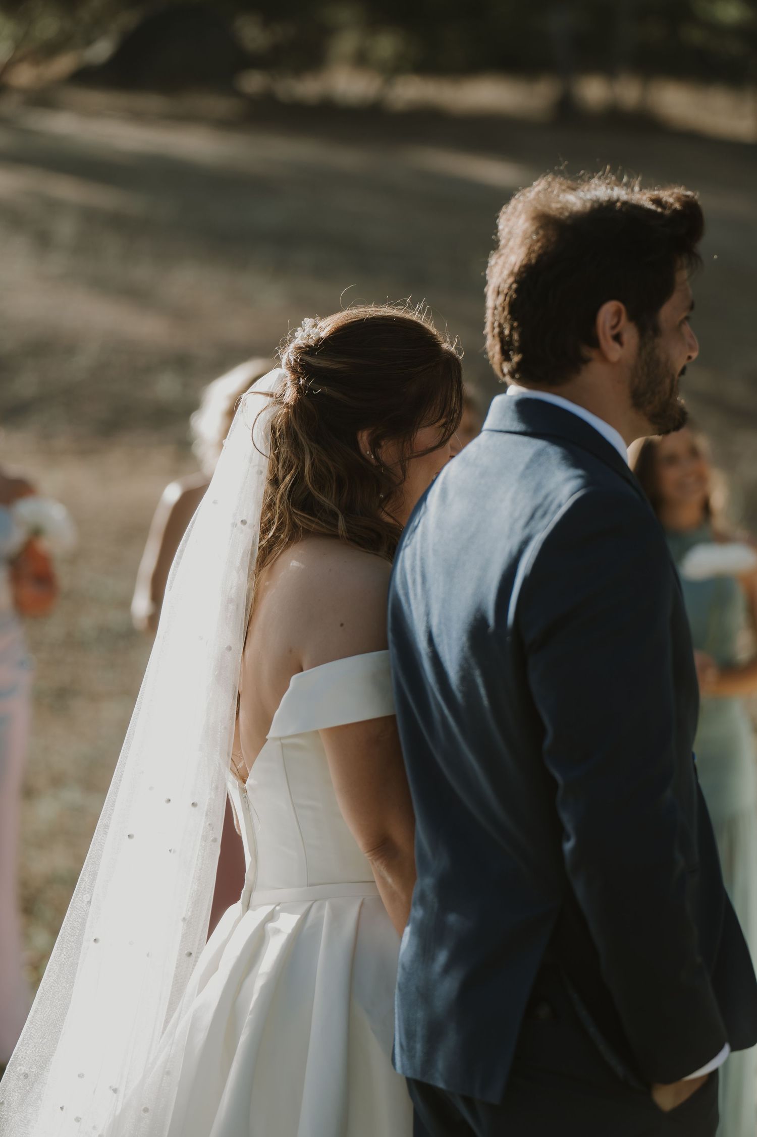 Bride in off-shoulder white gown with veil stands beside groom in navy suit at outdoor wedding ceremony.