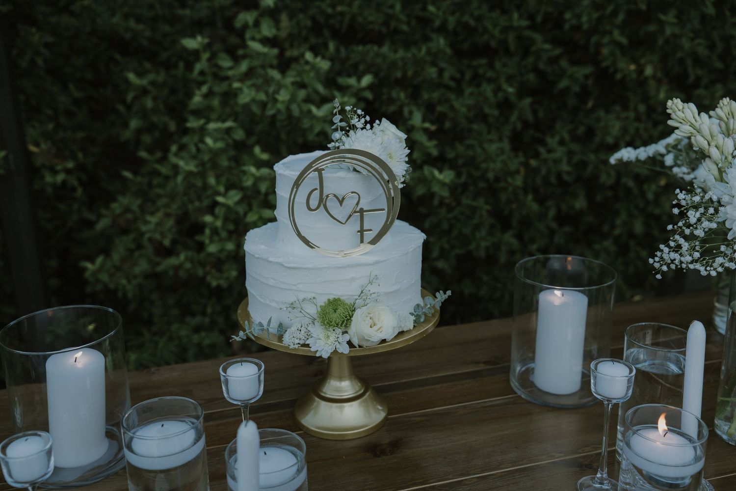 Two-tier white wedding cake with monogram topper on gold stand, surrounded by candles and greenery backdrop.