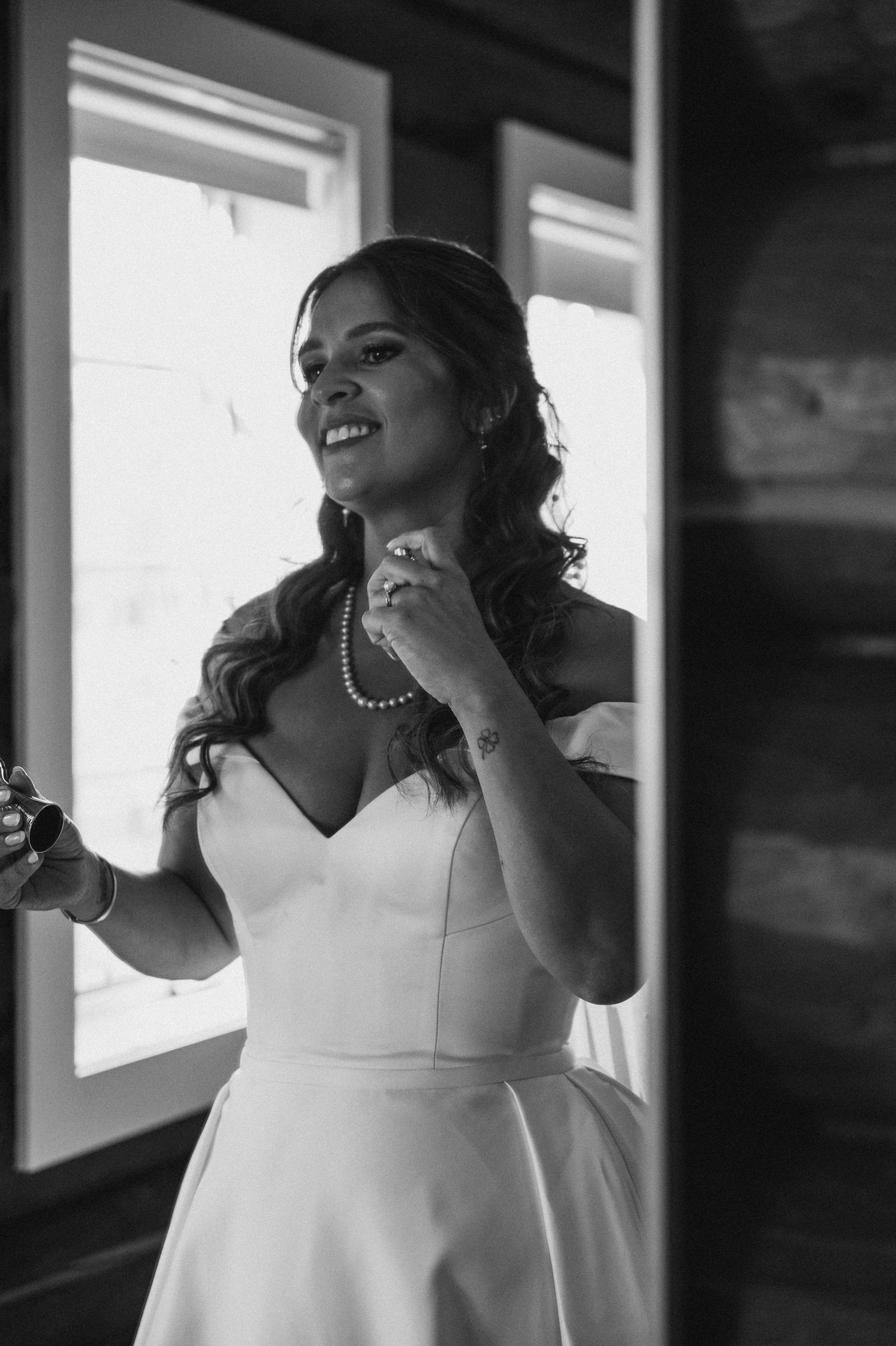 Smiling bride in off-shoulder white gown adjusting pearl necklace by window in black and white wedding photo.