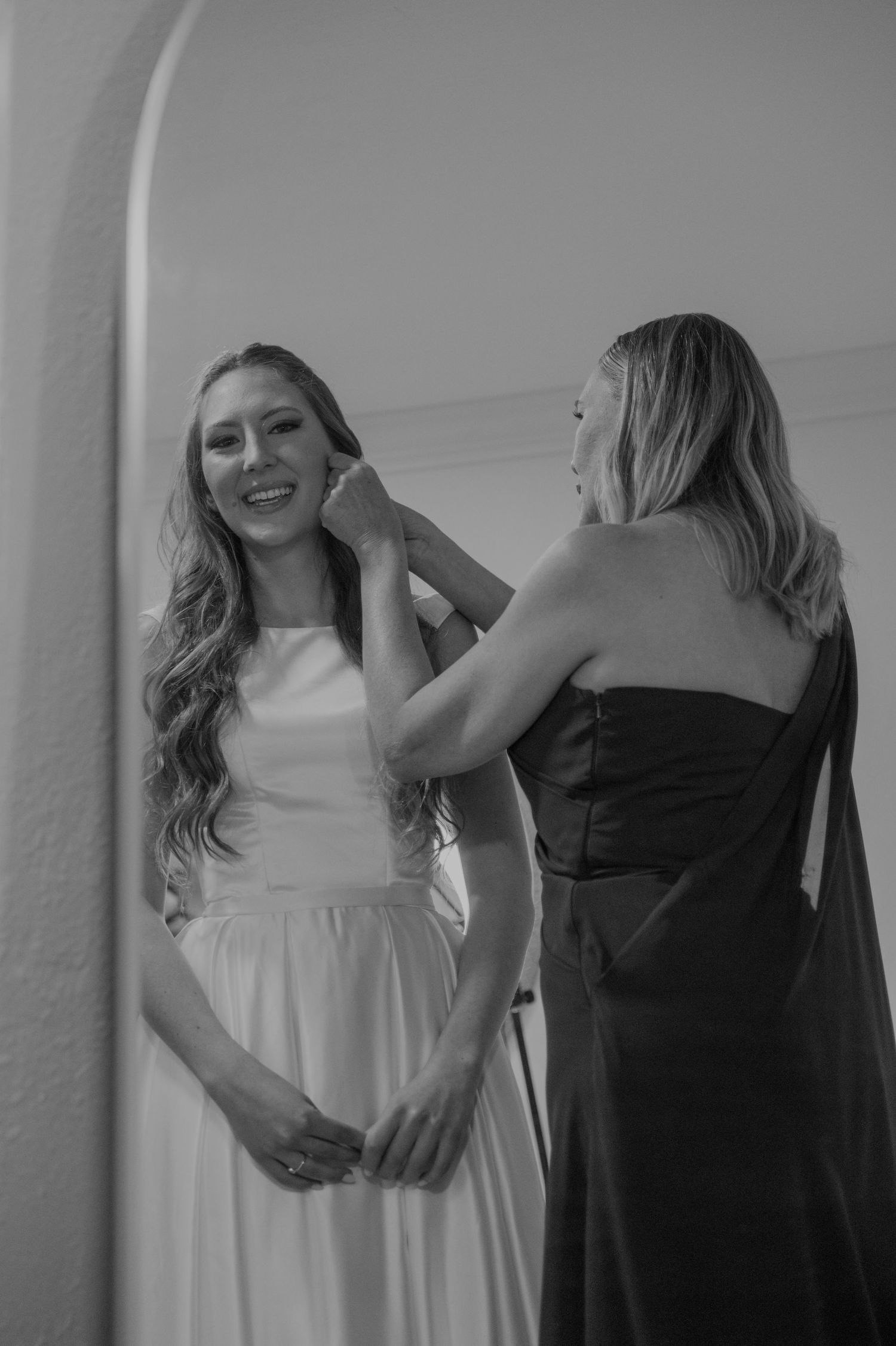 Black and white photo of smiling bride in wedding gown having earrings placed by a woman in black dress.
