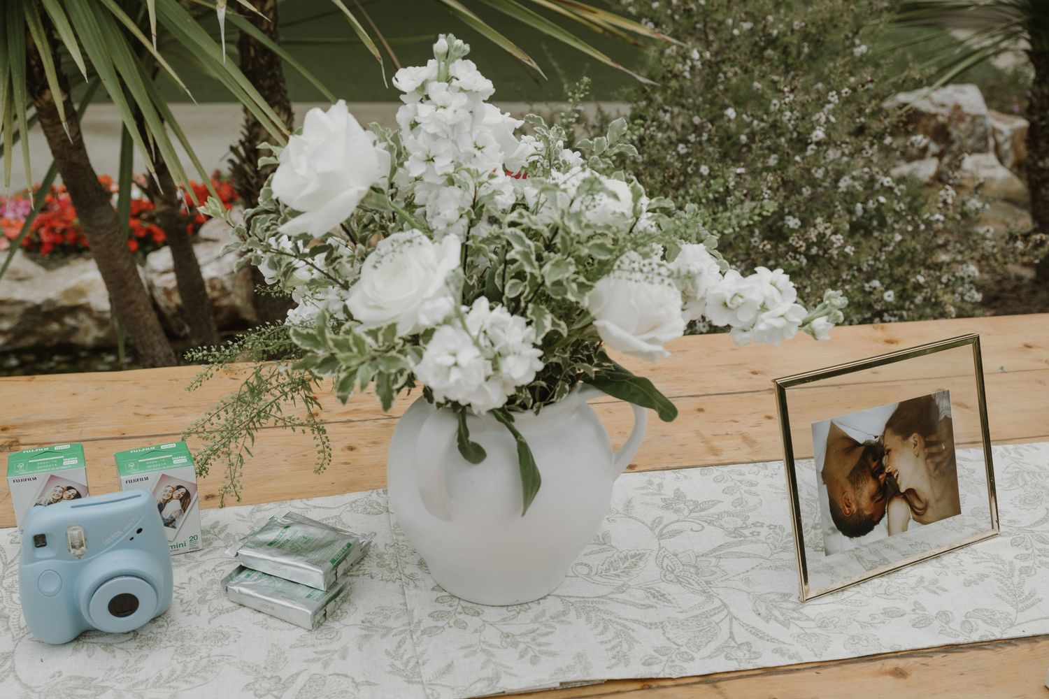 White peony floral arrangement in round vase beside Instax camera and framed photo on wedding table.