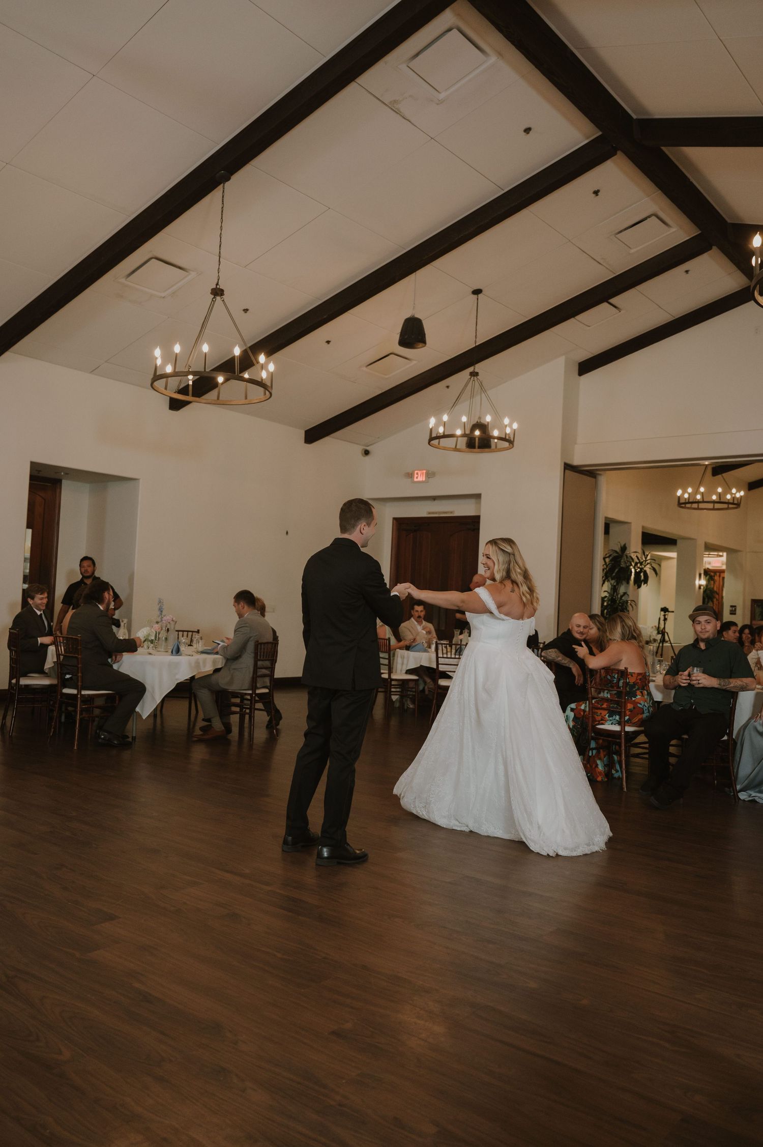Bride and groom sharing first dance in rustic ballroom with wood beam ceiling and chandeliers.