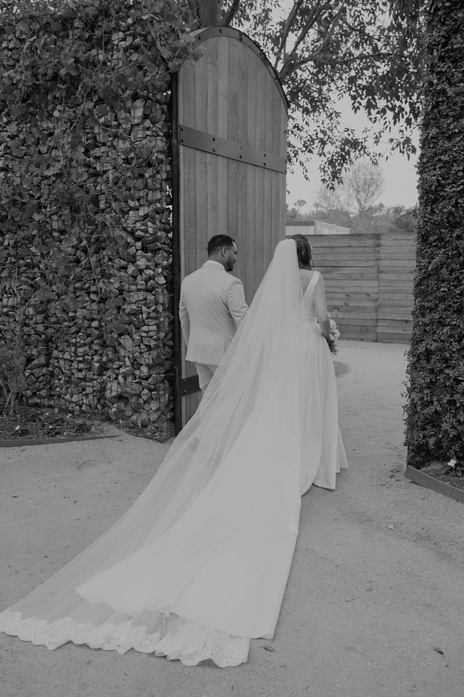 Black and white photo of bride and groom walking hand in hand through an arched wooden gate surrounded by ivy walls.