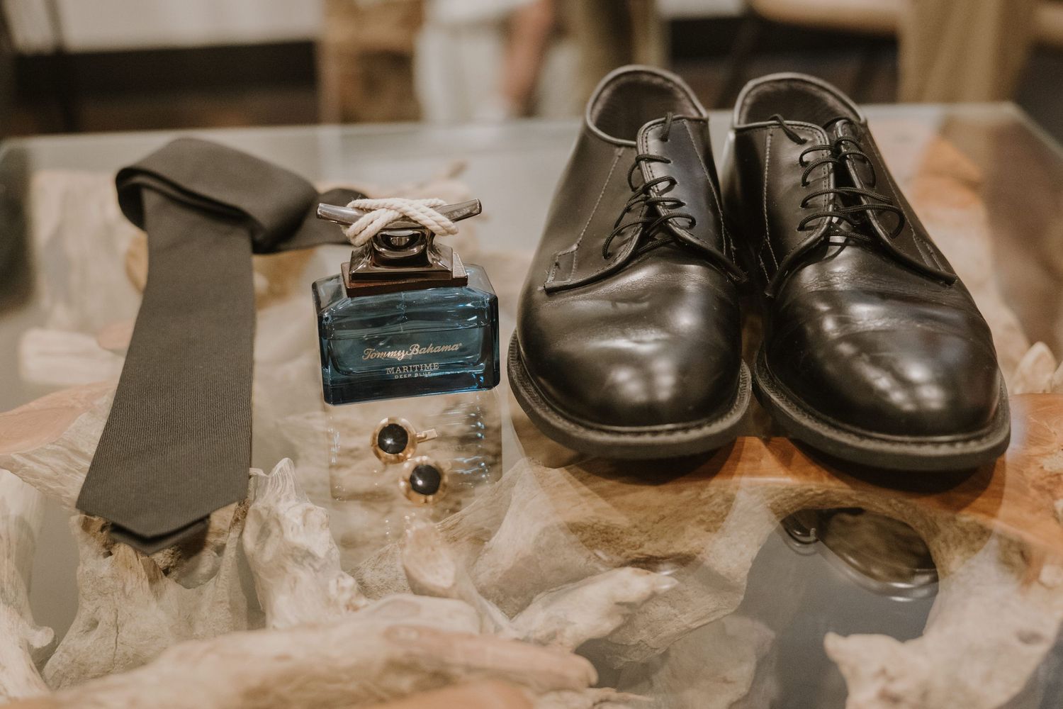 Black leather dress shoes, green tie, cologne bottle, and cufflinks displayed on a glass surface for a groom.