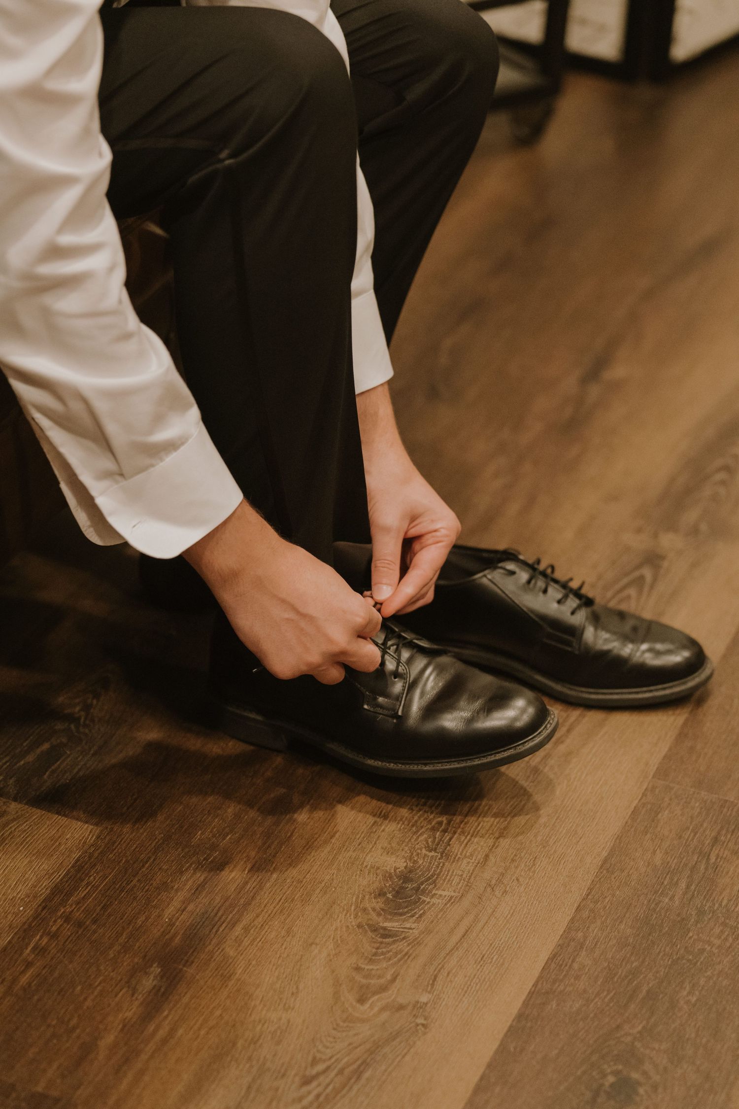 Man in white shirt and black pants tying laces on black oxford dress shoes on hardwood floor.
