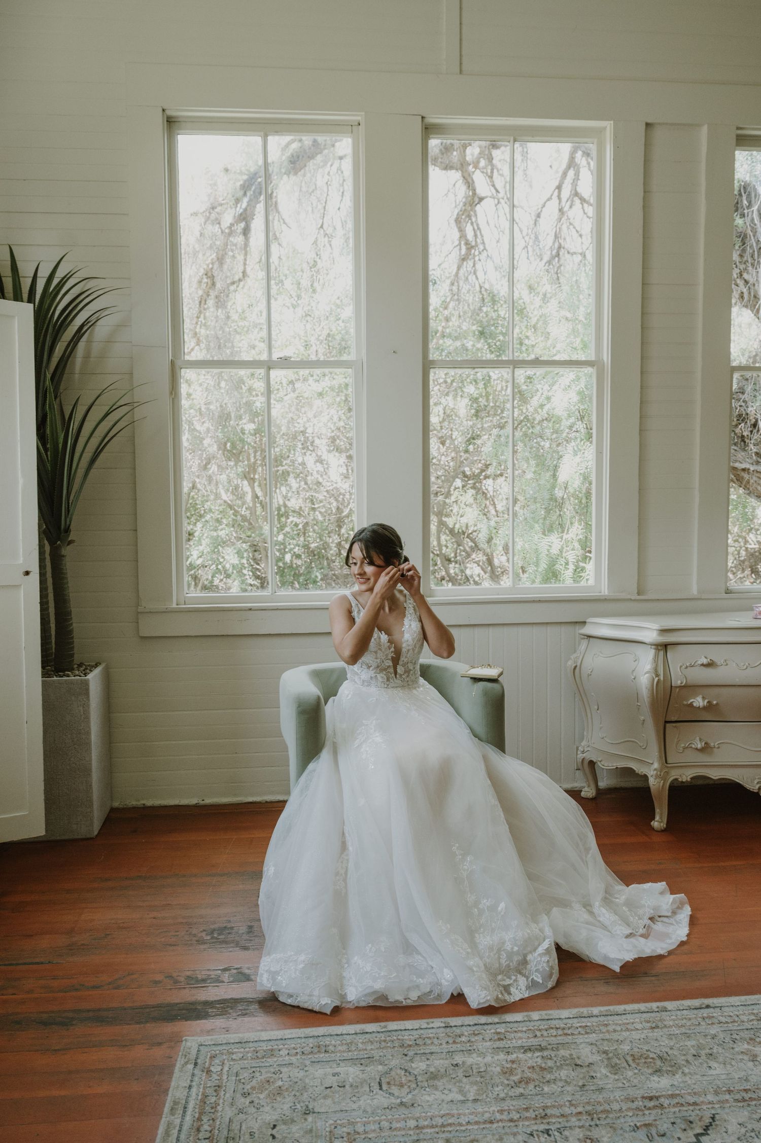 Bride in elegant lace ball gown sitting in a mint chair adjusting her veil by tall windows.
