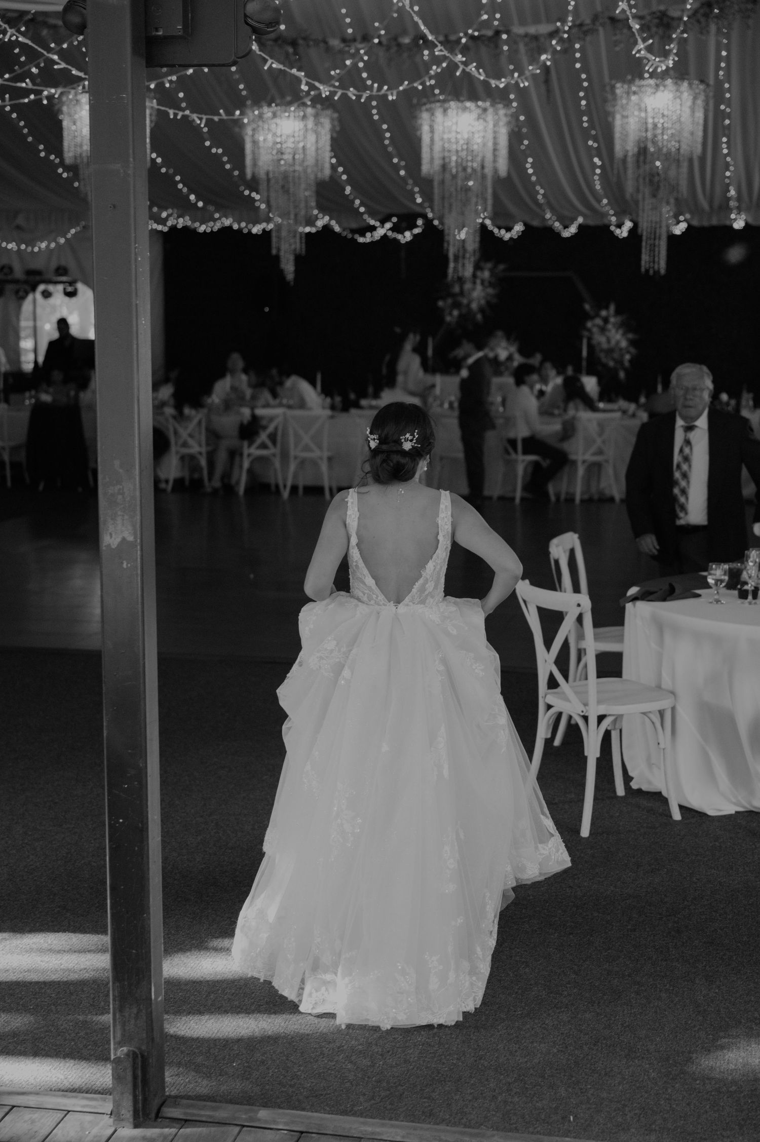 Bride in backless wedding gown entering elegant reception tent with fairy lights and chandeliers.