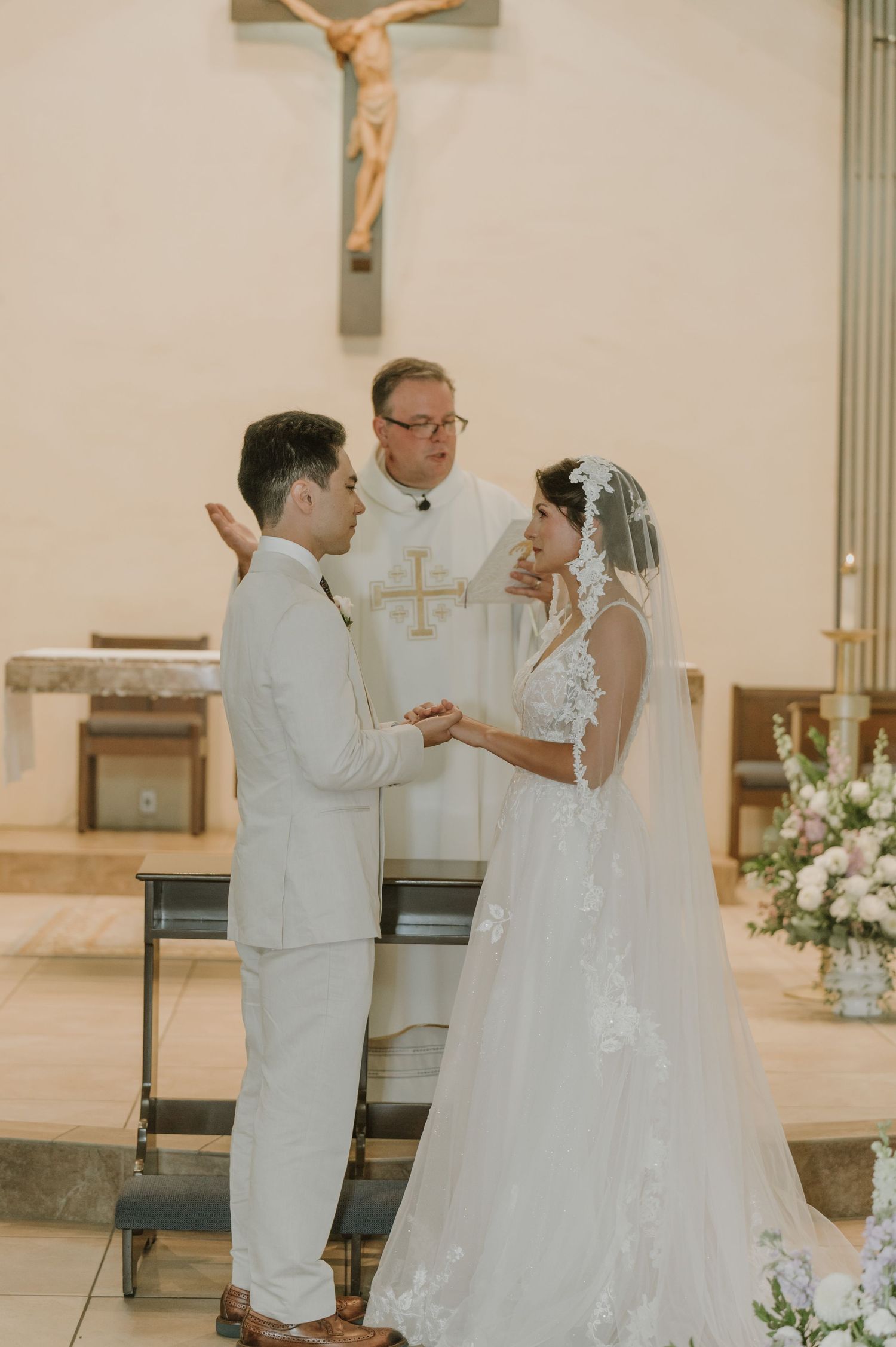 Bride and groom exchange vows before a priest beneath a crucifix in a Catholic church.