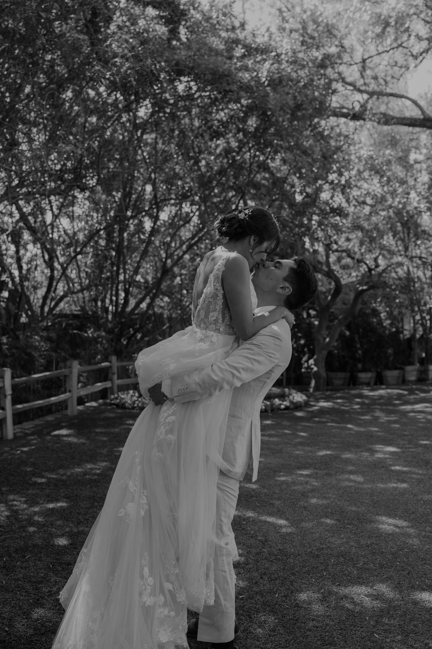 Groom lifts bride for a romantic kiss in a black and white outdoor wedding photo.