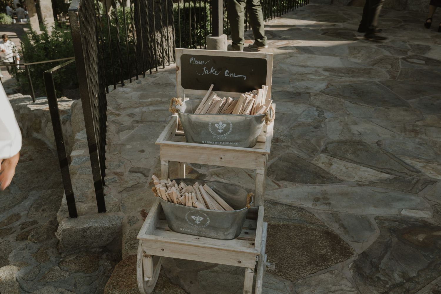 Rustic wooden cart display with books and programs in metal buckets at an outdoor wedding venue.
