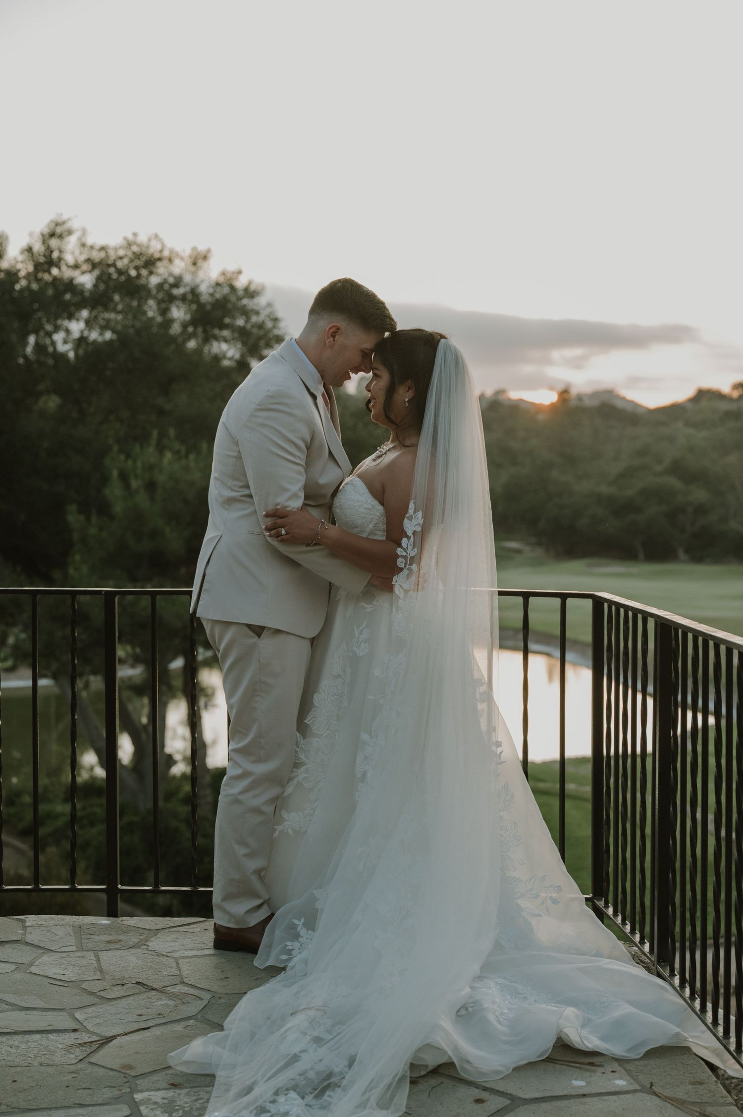 Bride and groom share a romantic kiss on a stone balcony at golden hour sunset.