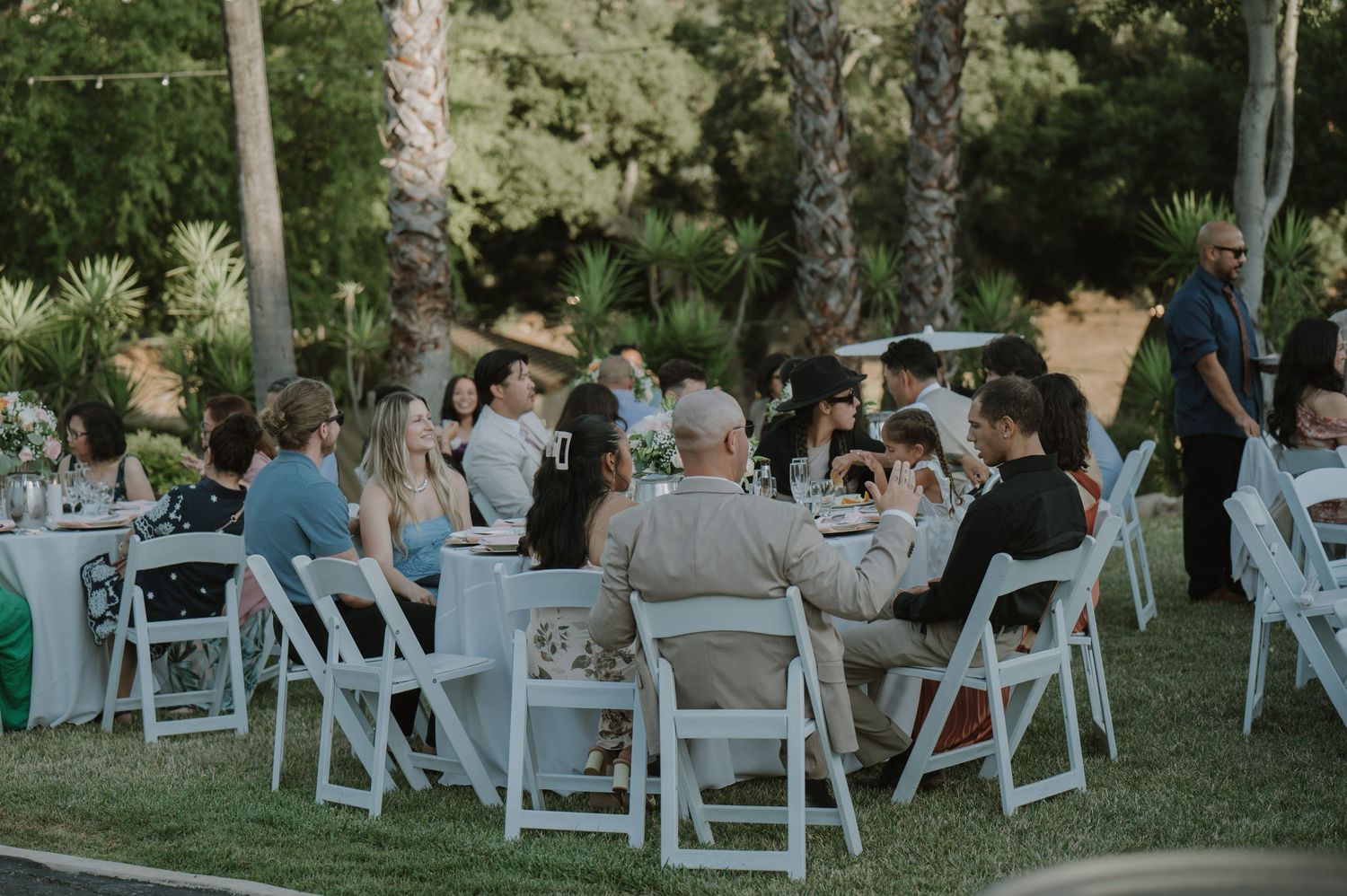 Guests seated at white outdoor tables on green lawn during an elegant al fresco wedding reception.