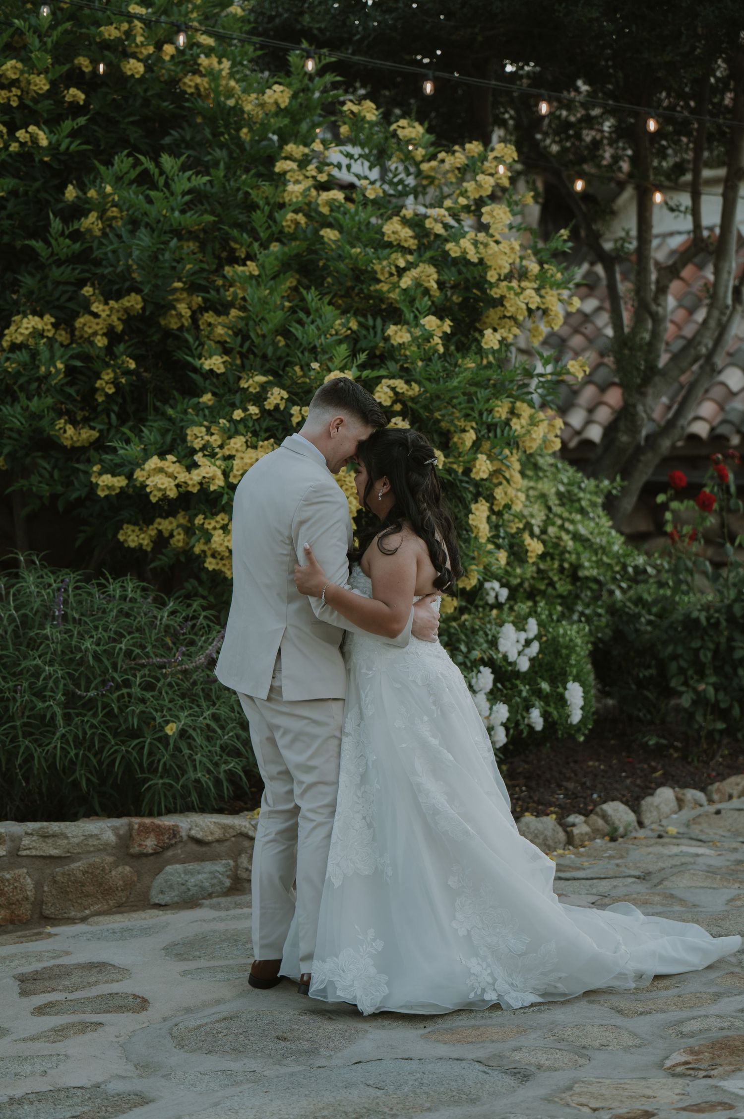 Bride and groom embrace tenderly in a lush garden with yellow blooms and string lights at their outdoor wedding.