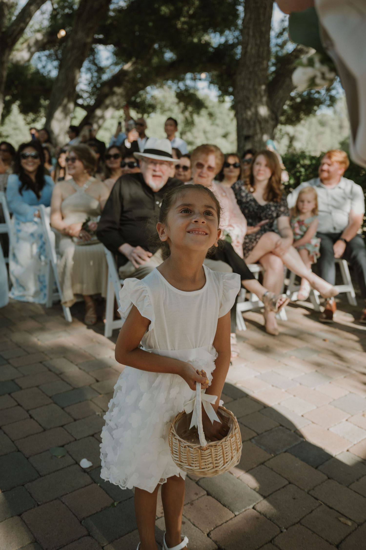 Young flower girl in white dress carries wicker basket down sunny outdoor wedding ceremony aisle.