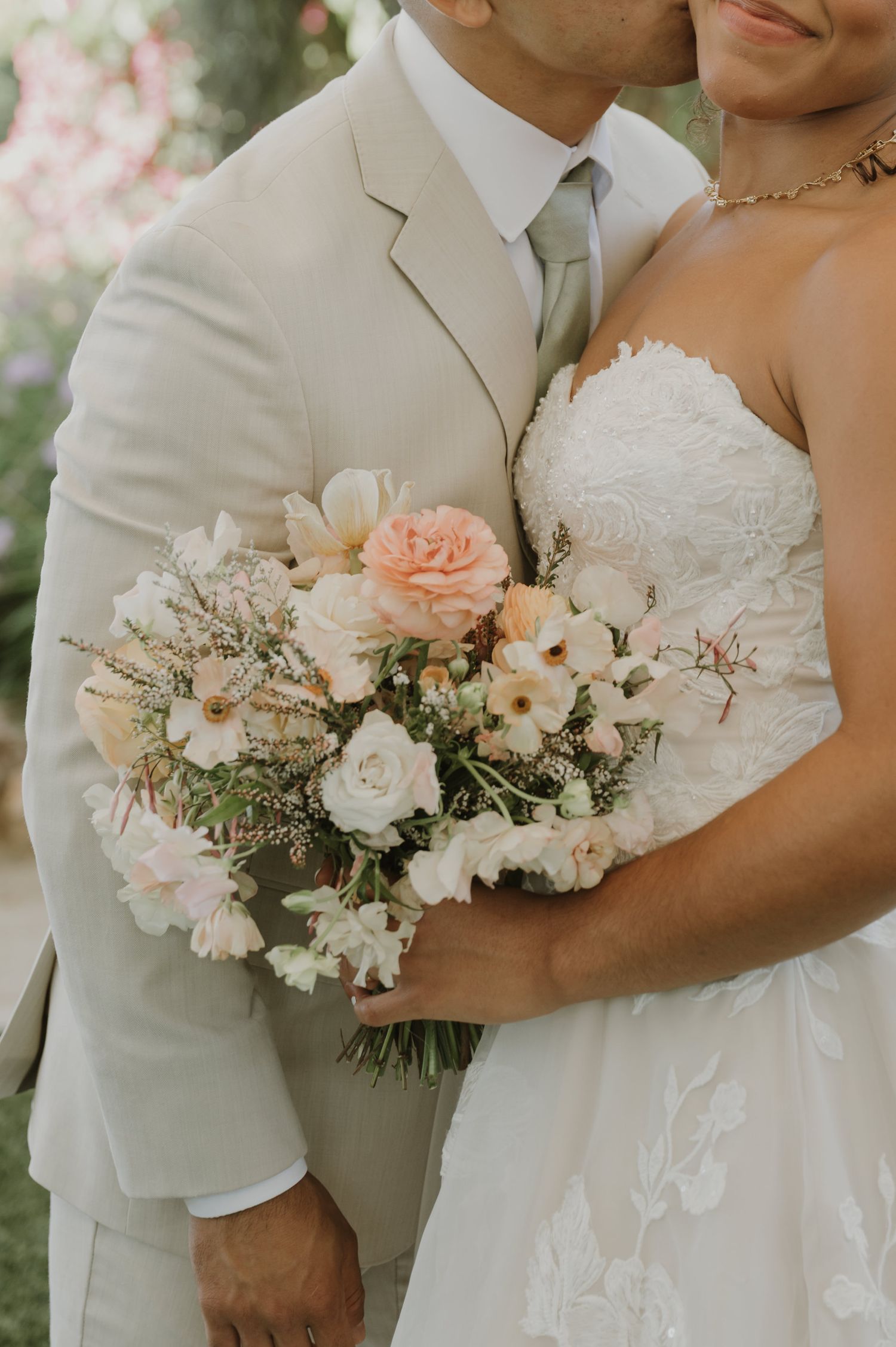 Bride in strapless lace gown holds soft pink and white bouquet while groom in tan suit embraces her outdoors.