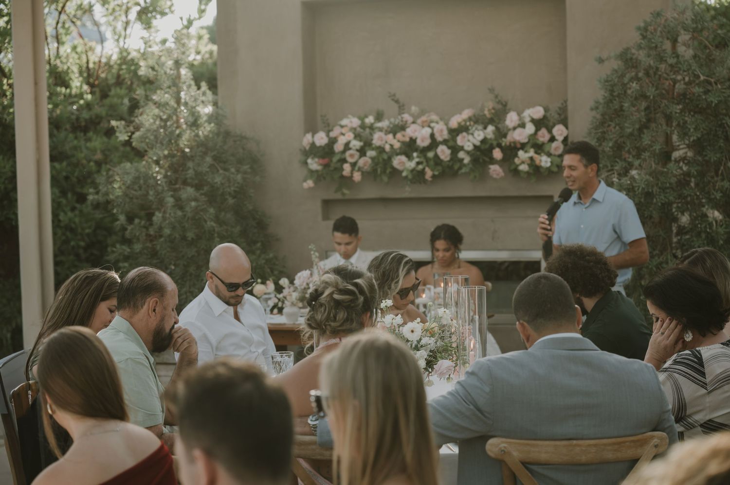 Wedding guests seated at an outdoor reception dinner while a man gives a heartfelt speech.