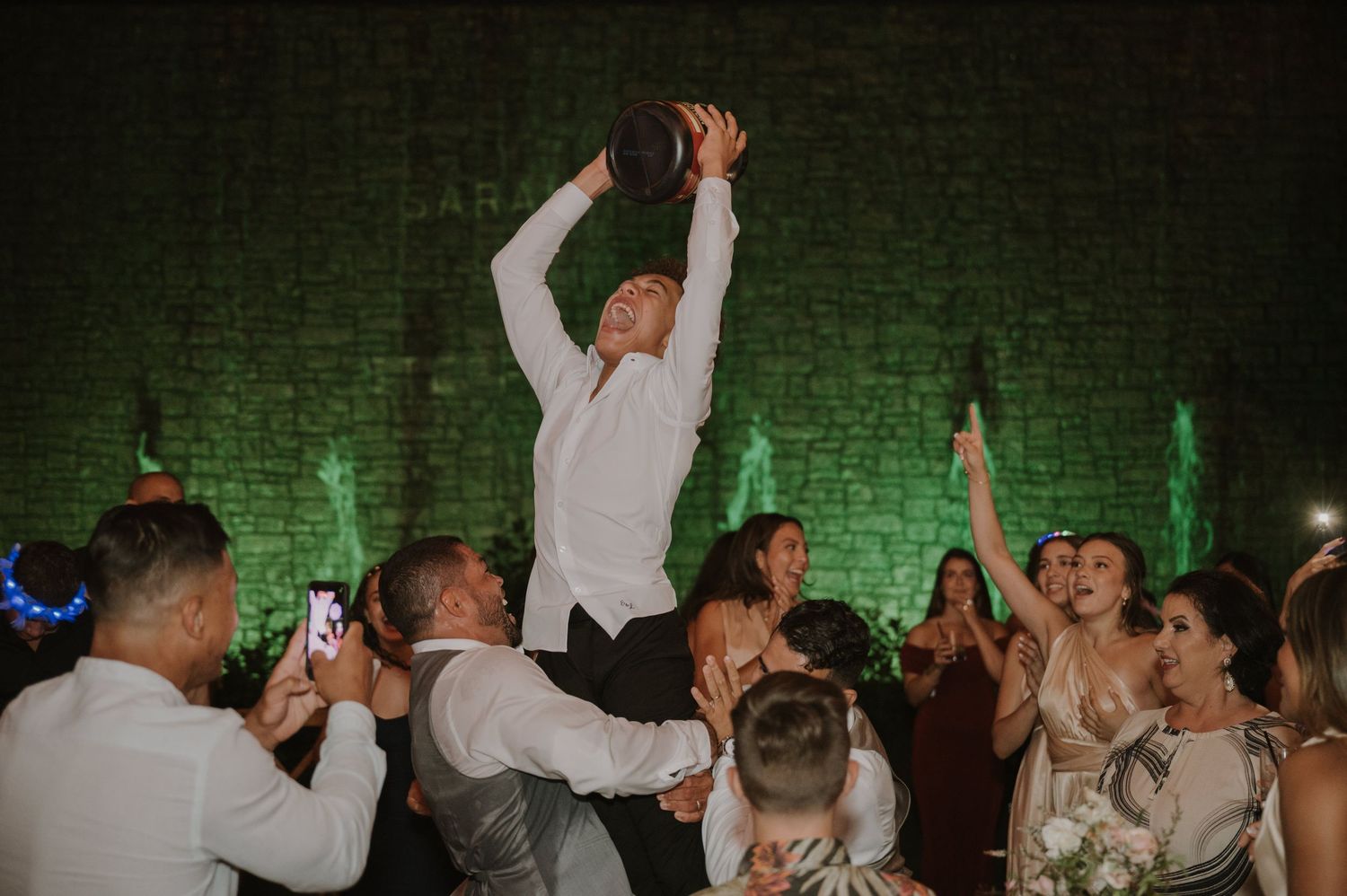 Groom lifted by guests holding bouquet aloft during lively wedding reception dance celebration.
