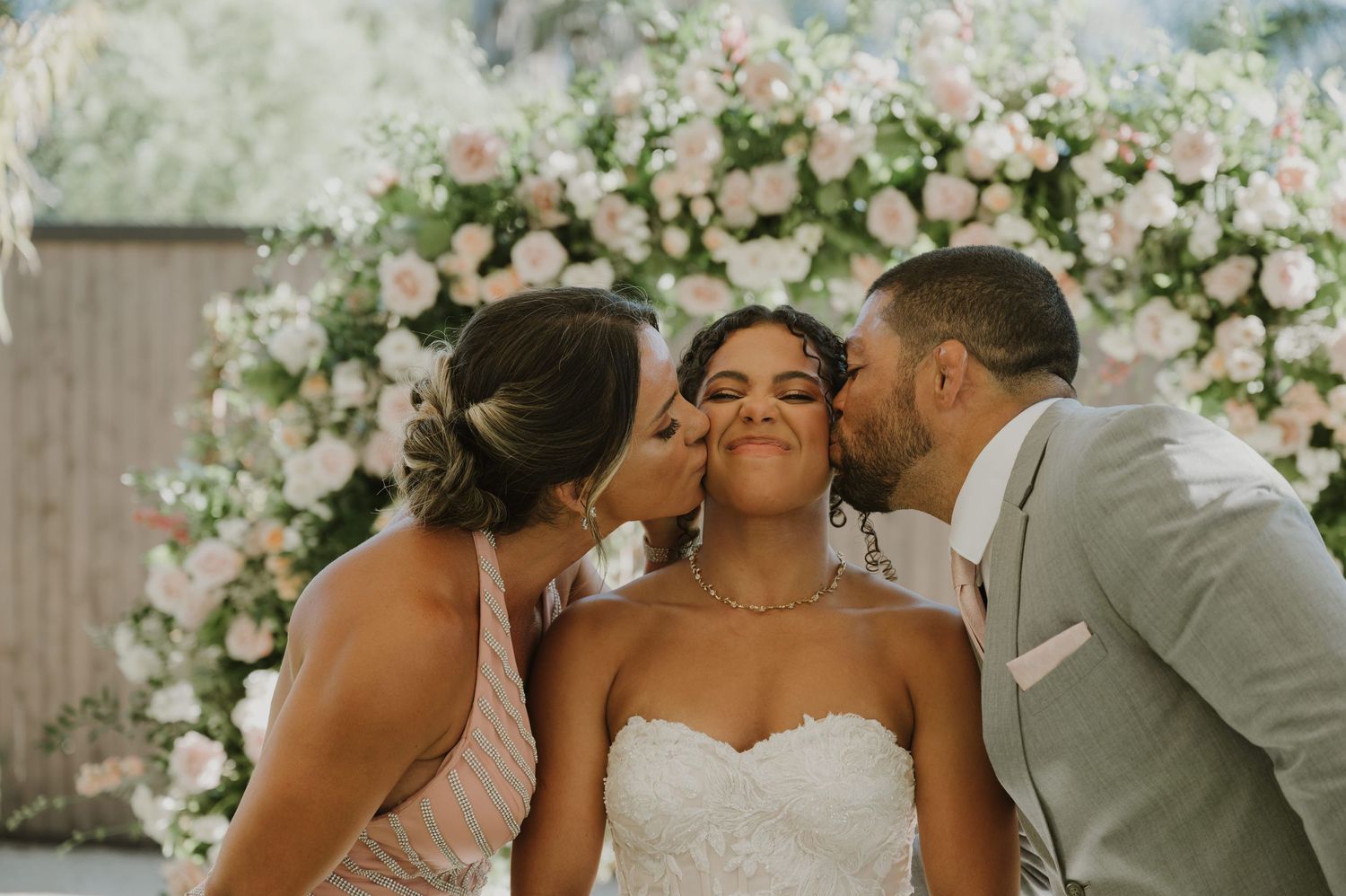 Bride receiving kisses on cheeks from bridesmaid and groom in front of pink floral arch.