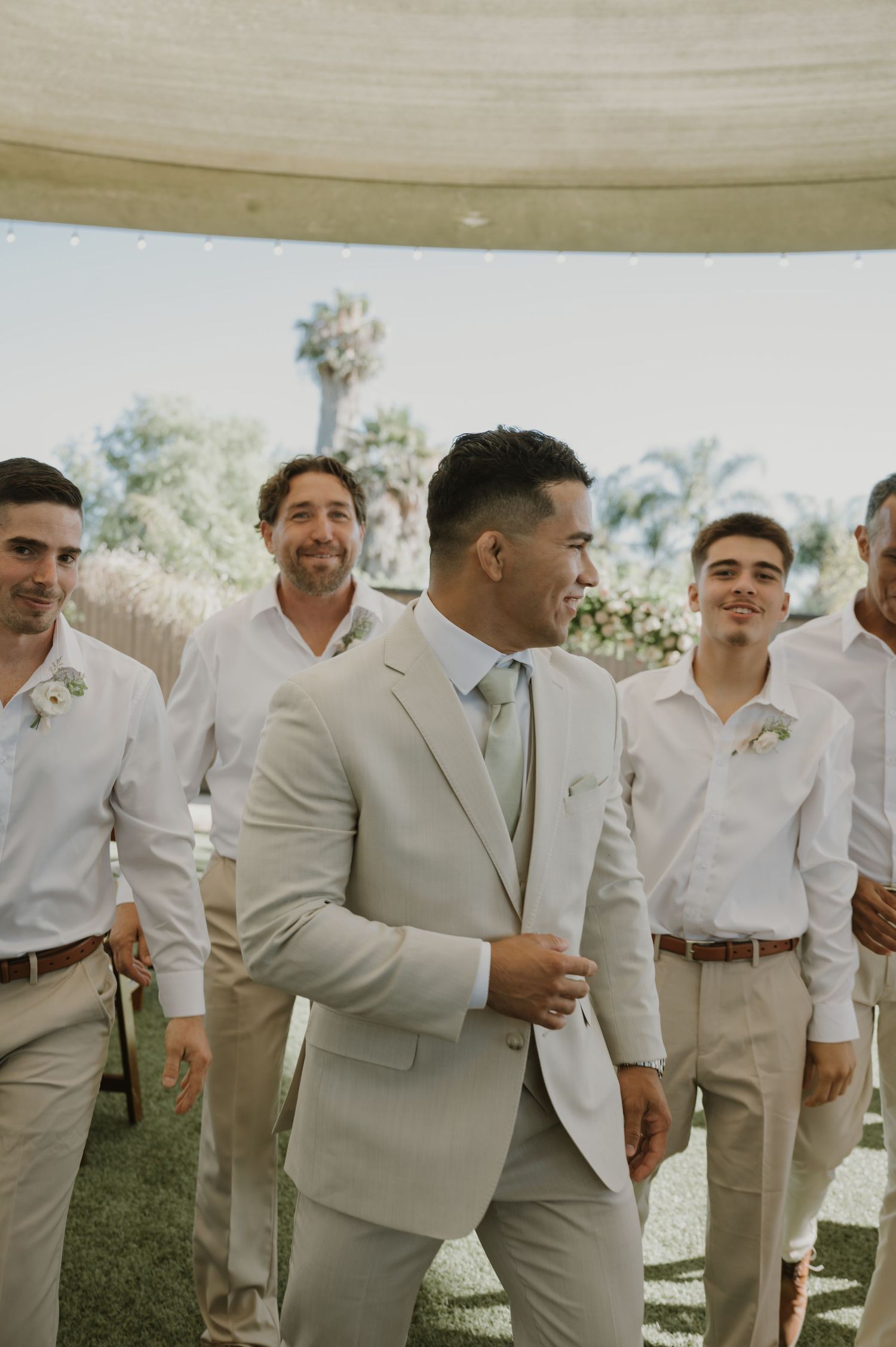 Groom in beige suit smiles with groomsmen in white shirts at outdoor wedding ceremony.