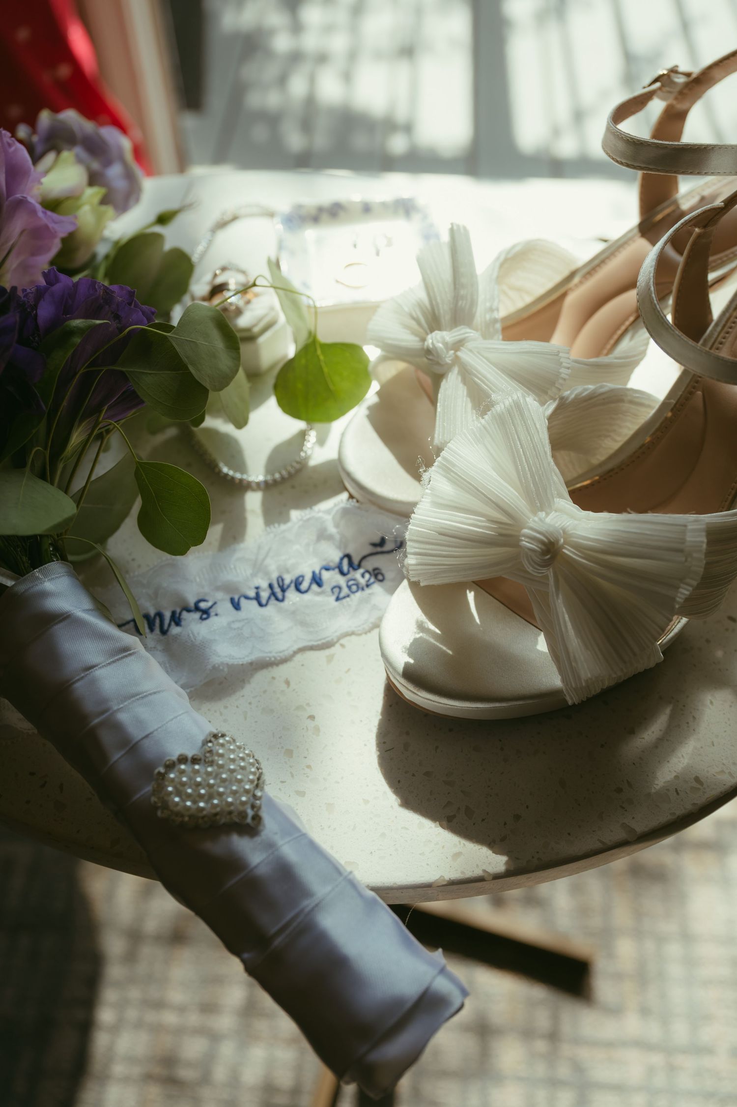 Elegant bridal flat lay featuring white bow heels, wedding bouquet, Mrs. Rivera garter, pearl brooch, and gold rings on a marble surface.
