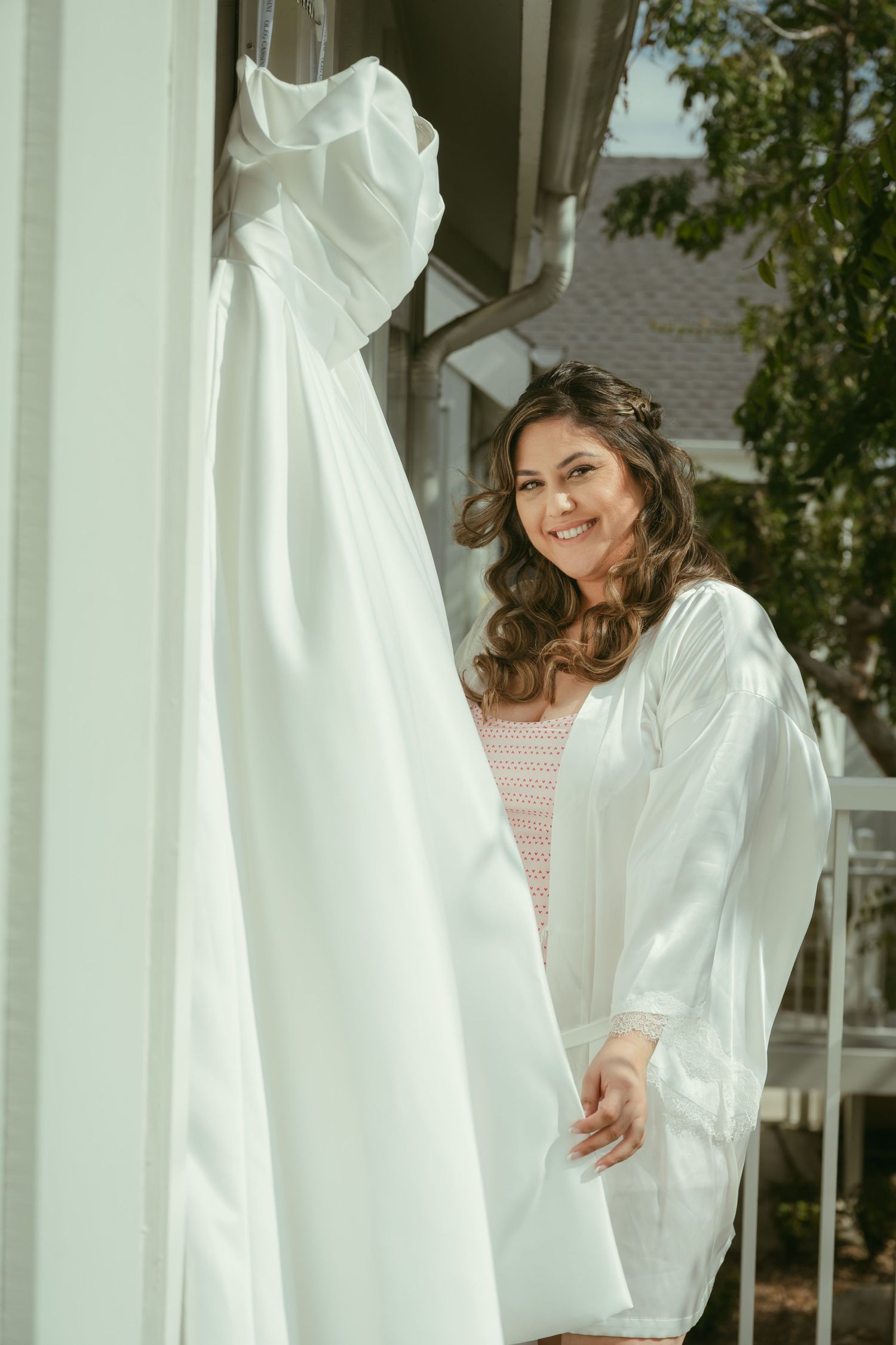 Smiling bride in white robe standing beside her elegant wedding gown hanging outdoors before ceremony.