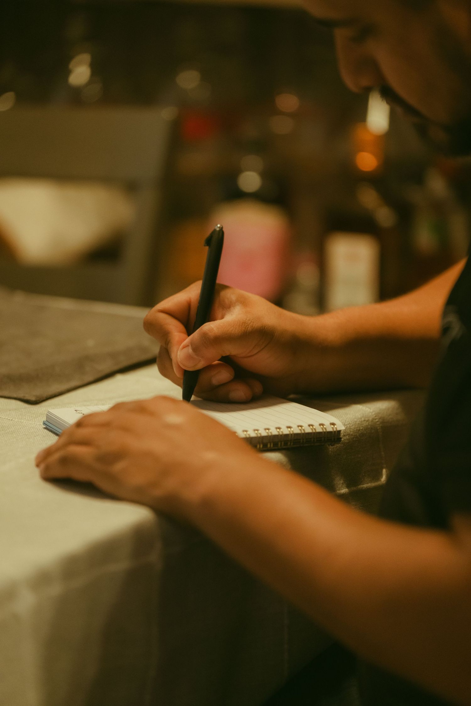 A person writing notes in a spiral notebook with a black pen at a table in a warmly lit restaurant.