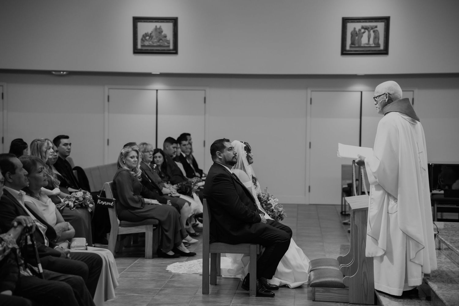 Black and white wedding ceremony photo with couple seated before priest at altar, guests watching in church pews.