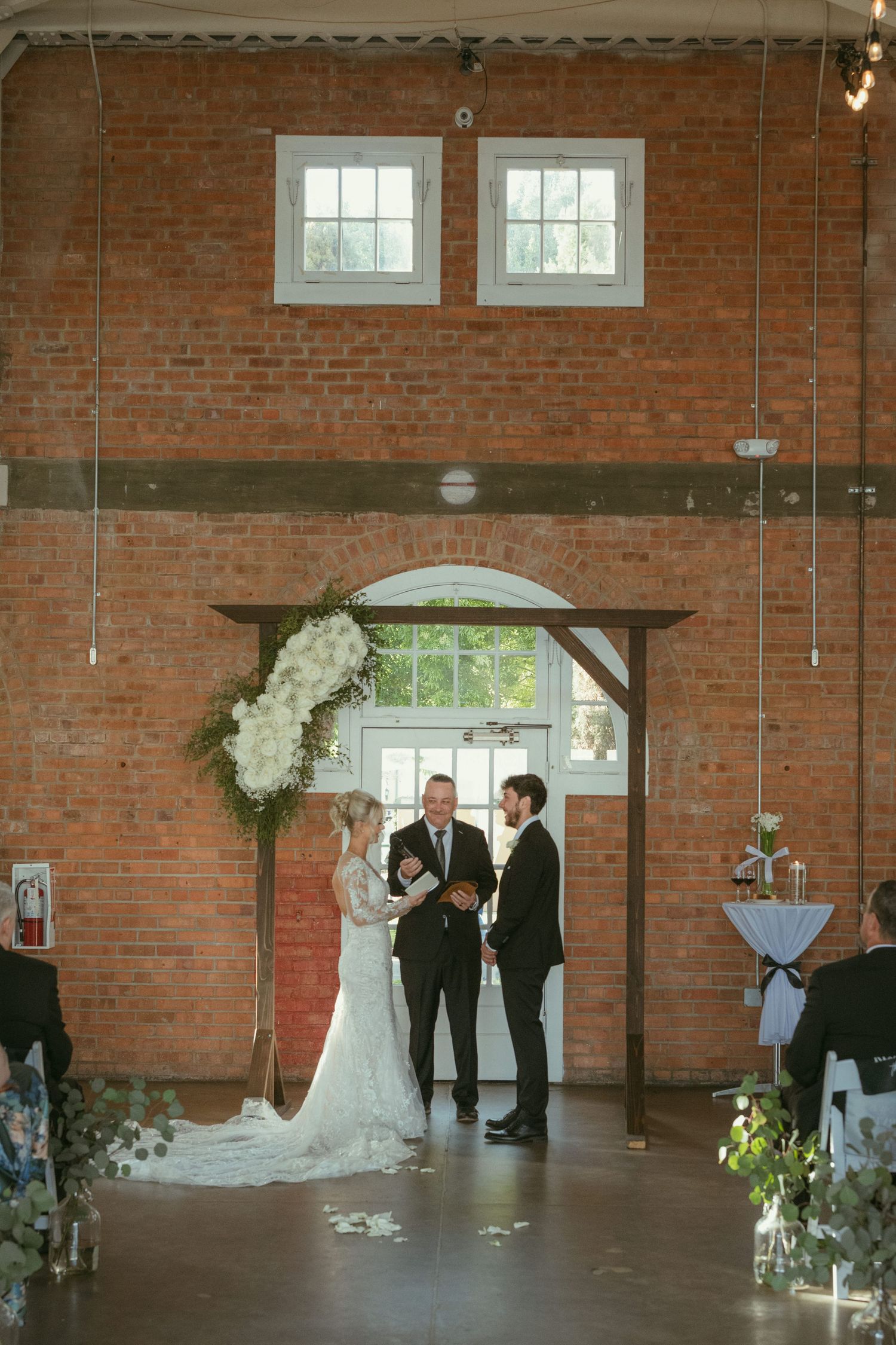 Bride and groom exchange vows under floral arch in rustic brick venue with officiant during indoor wedding ceremony.