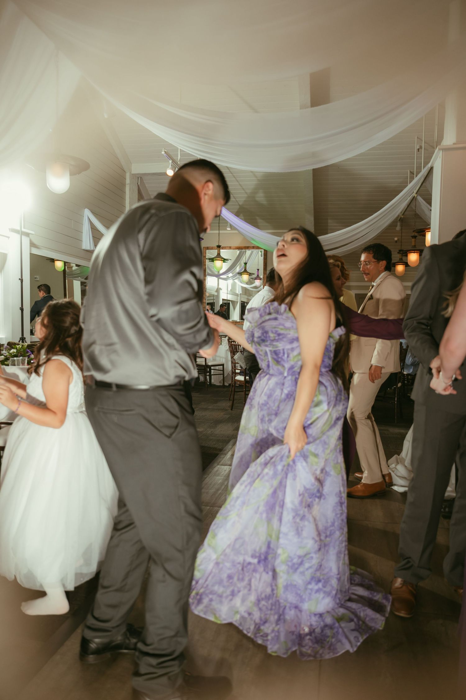 Wedding guest in purple floral gown dancing energetically with partner on decorated reception dance floor.