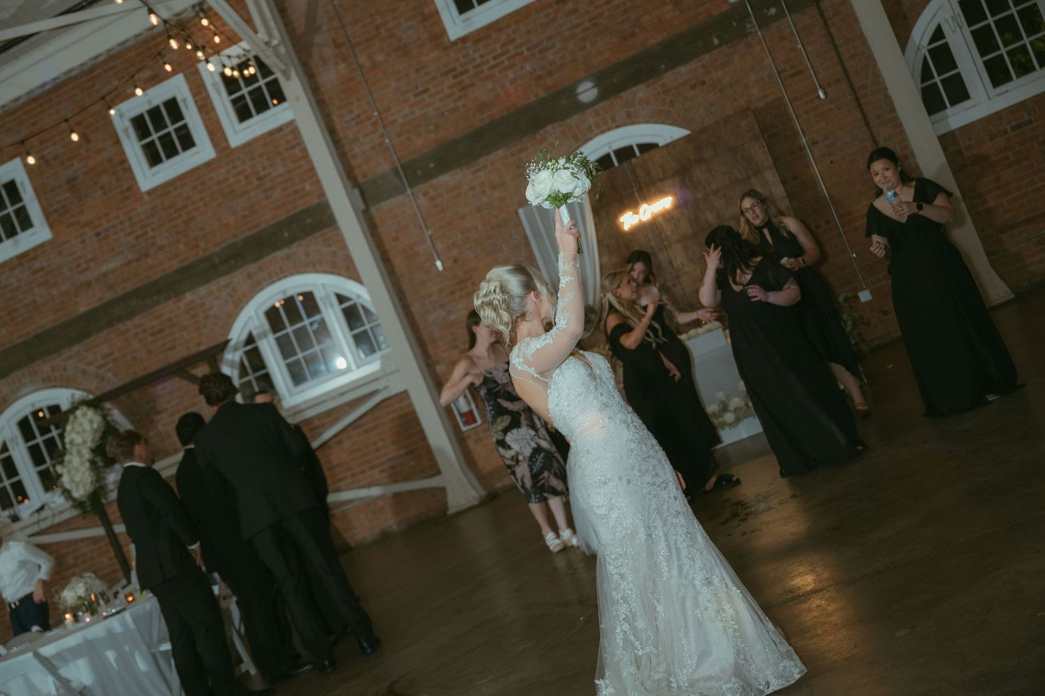 Bride in white lace gown tosses her bouquet to excited guests at wedding reception.