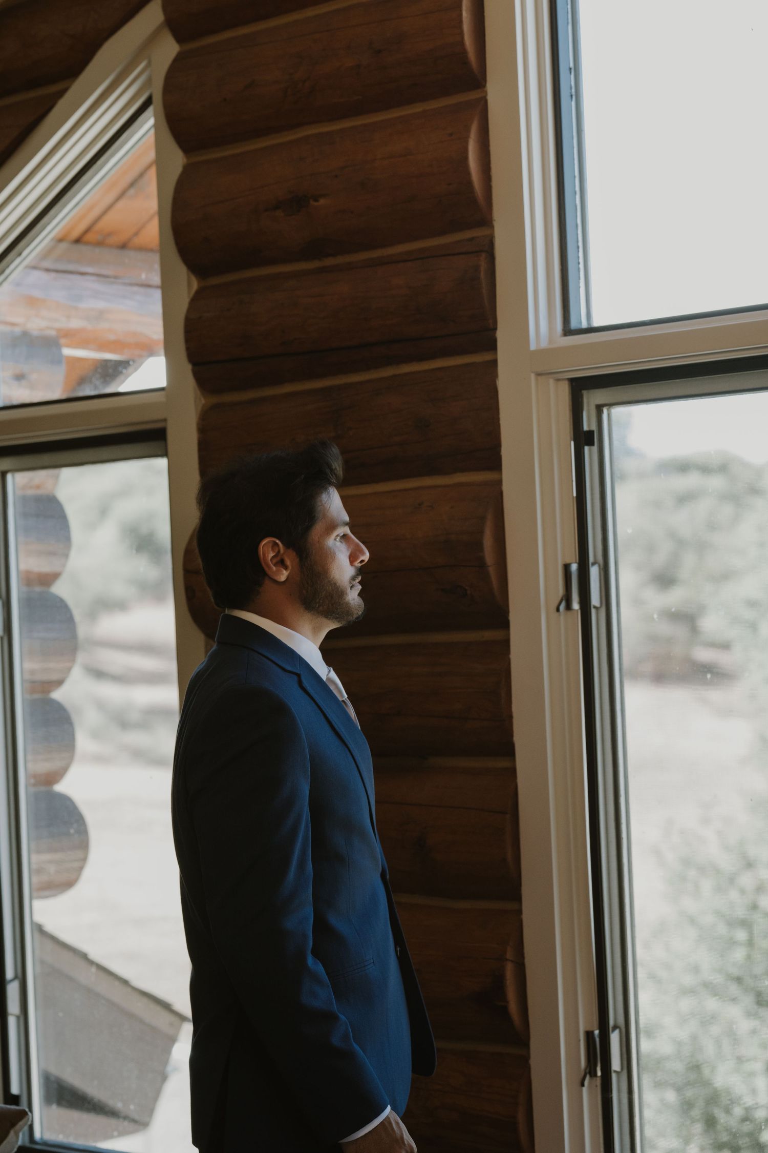 Groom in navy suit stands by large windows of rustic log cabin, gazing outside on wedding day.