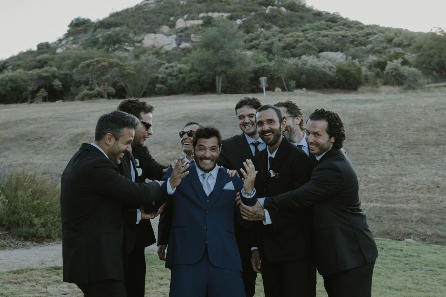 Groomsmen in black suits surround and celebrate the groom in a blue suit on a scenic hillside.