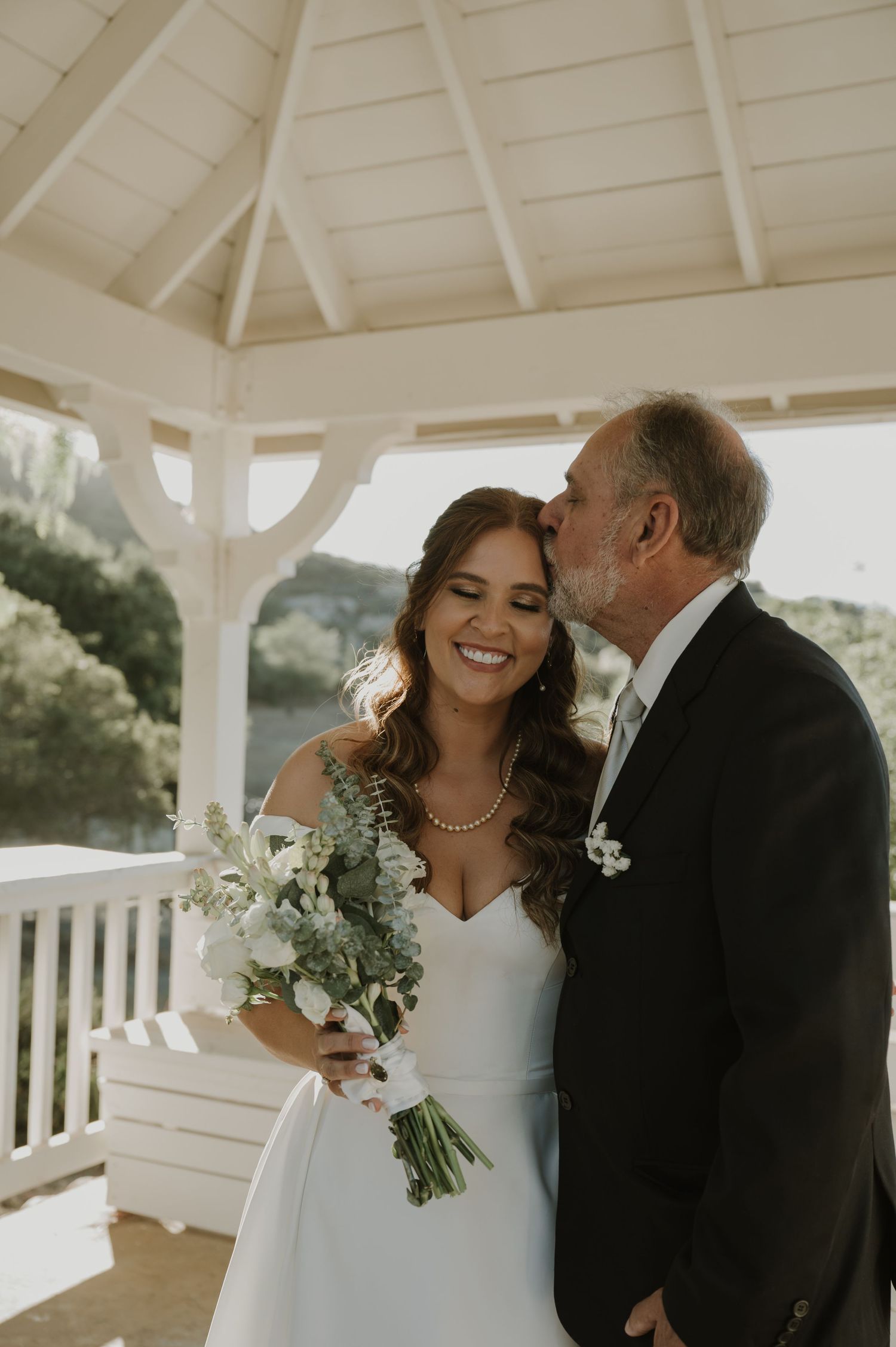 Bride smiling as older man kisses her cheek while she holds white floral bouquet in gazebo.