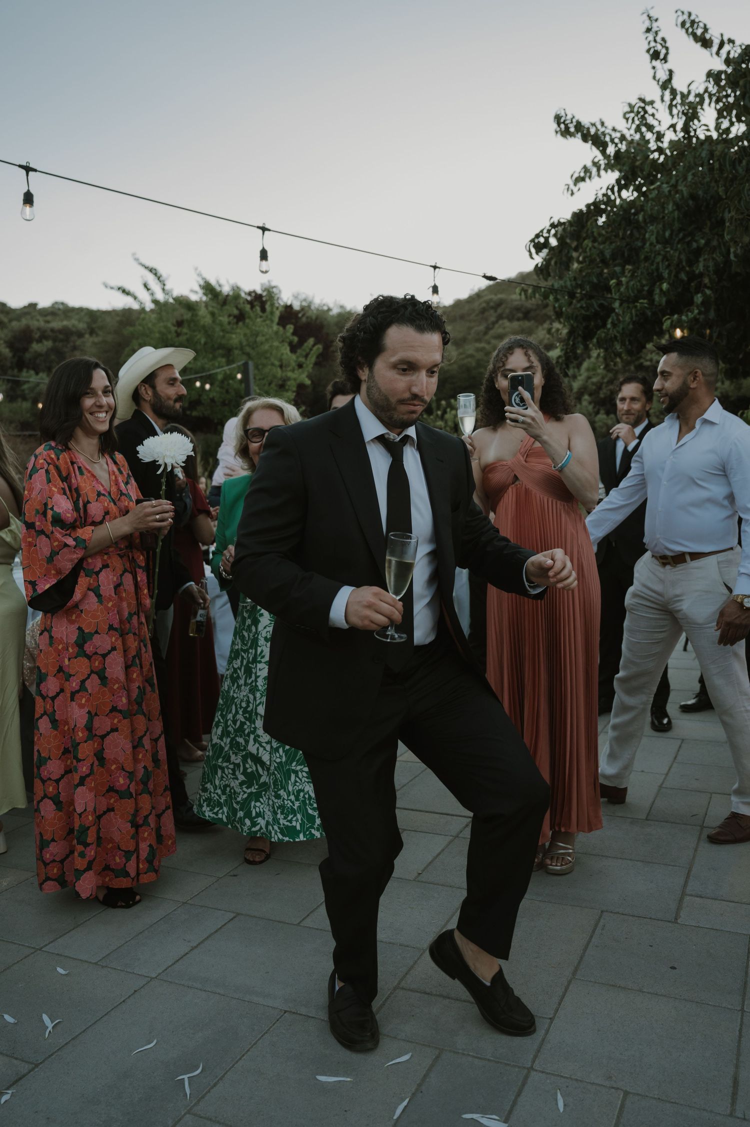 Man in black suit dancing at outdoor wedding reception while guests watch and smile.