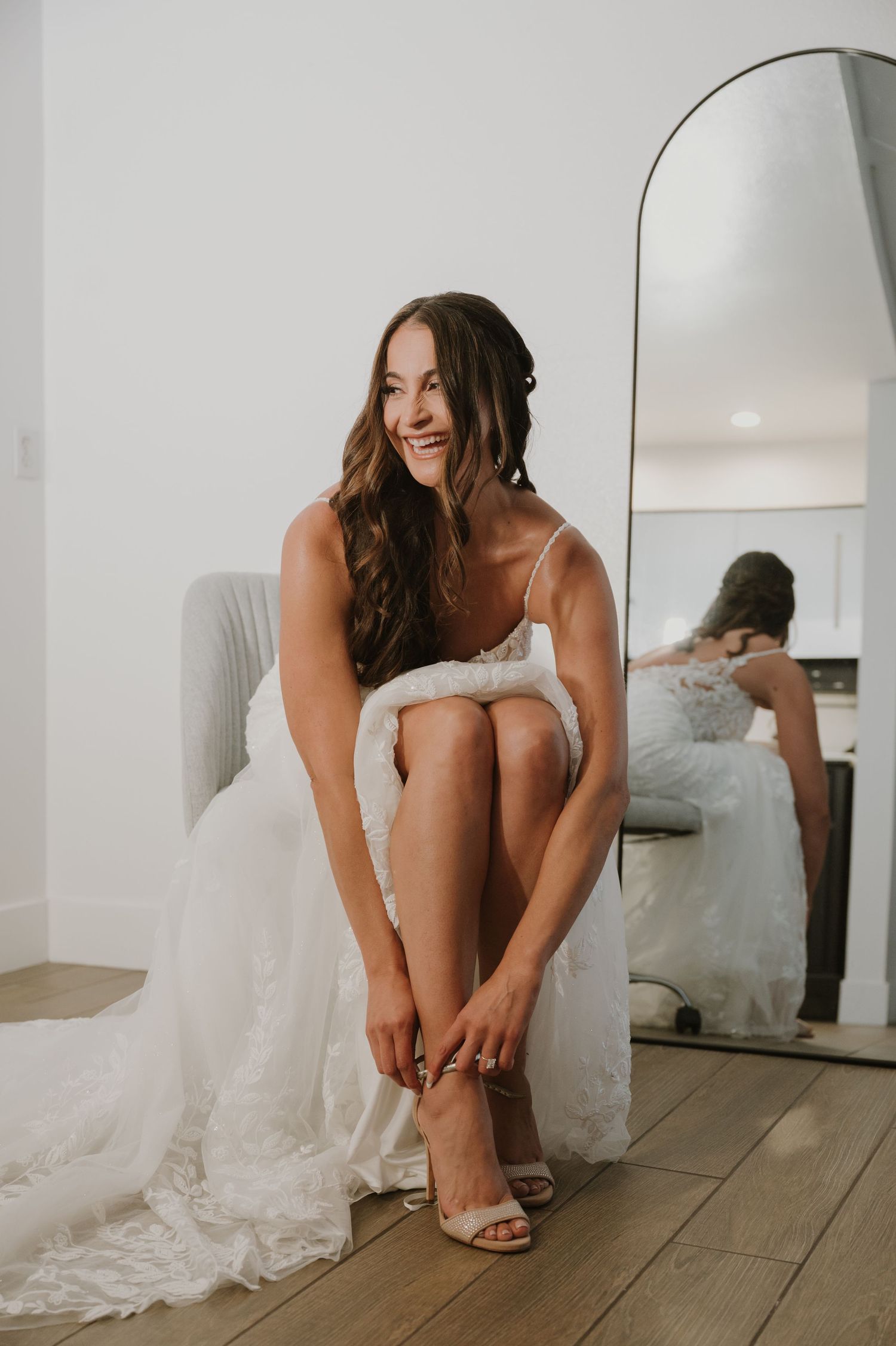 Bride in white lace wedding dress adjusting her heel sandals while seated, smiling near a full-length mirror.