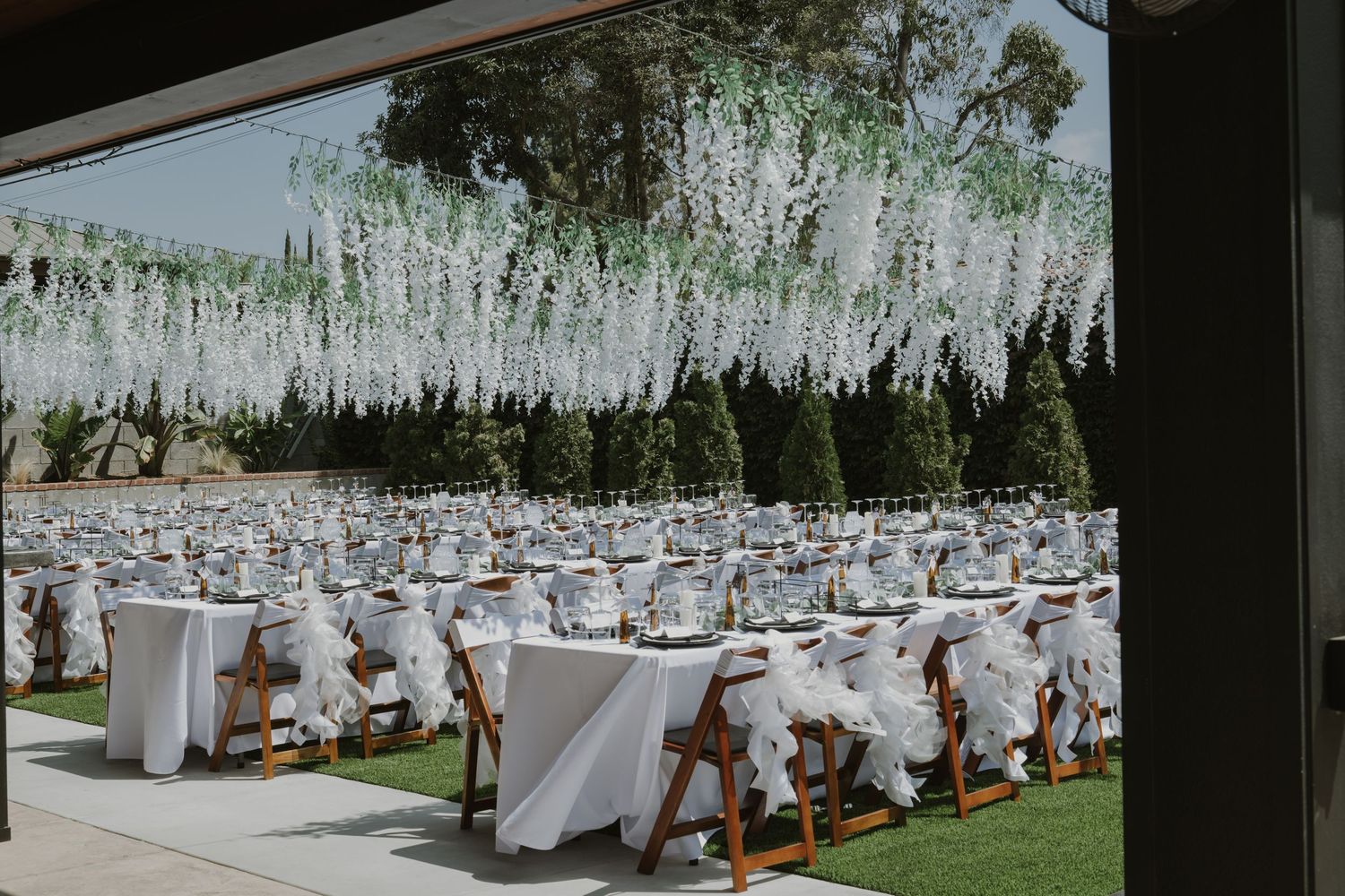 Elegant outdoor wedding reception with white draped tables, wooden chairs, and hanging floral arrangements overhead.