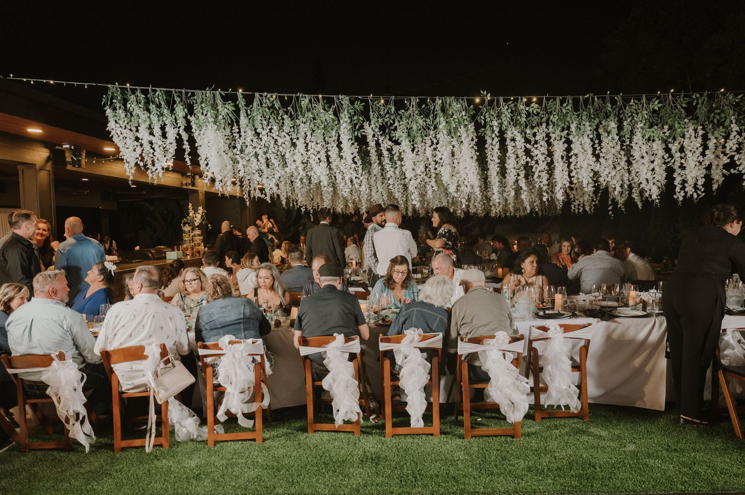 Guests dining at outdoor wedding reception under hanging white floral canopy at night.