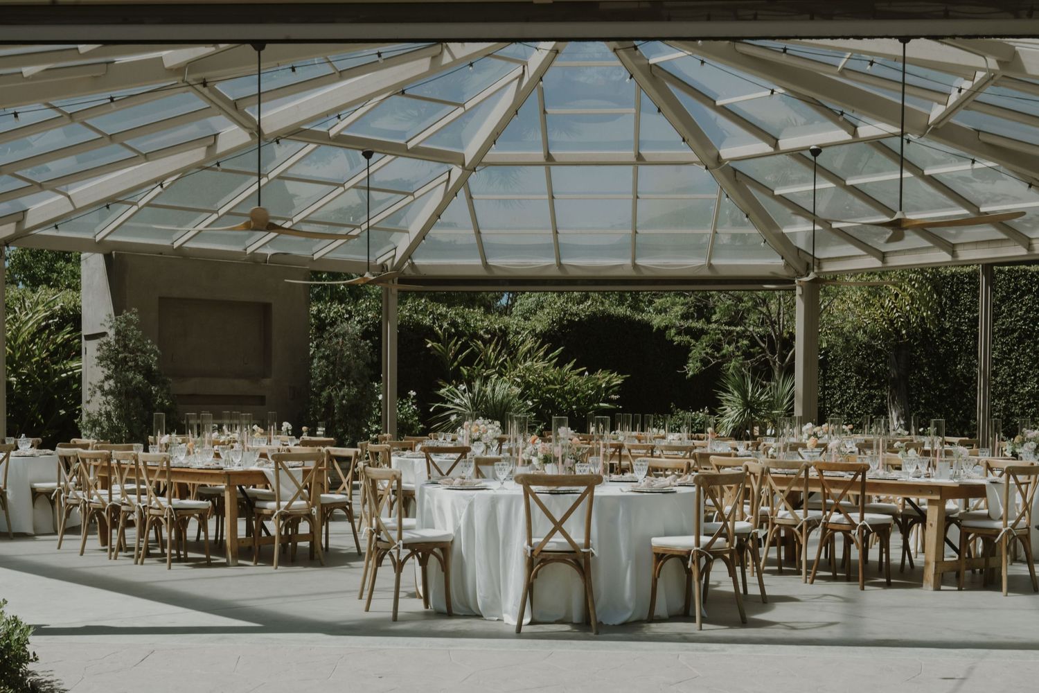 Elegant wedding reception under a glass conservatory roof with round tables, white linens, and wooden cross-back chairs.