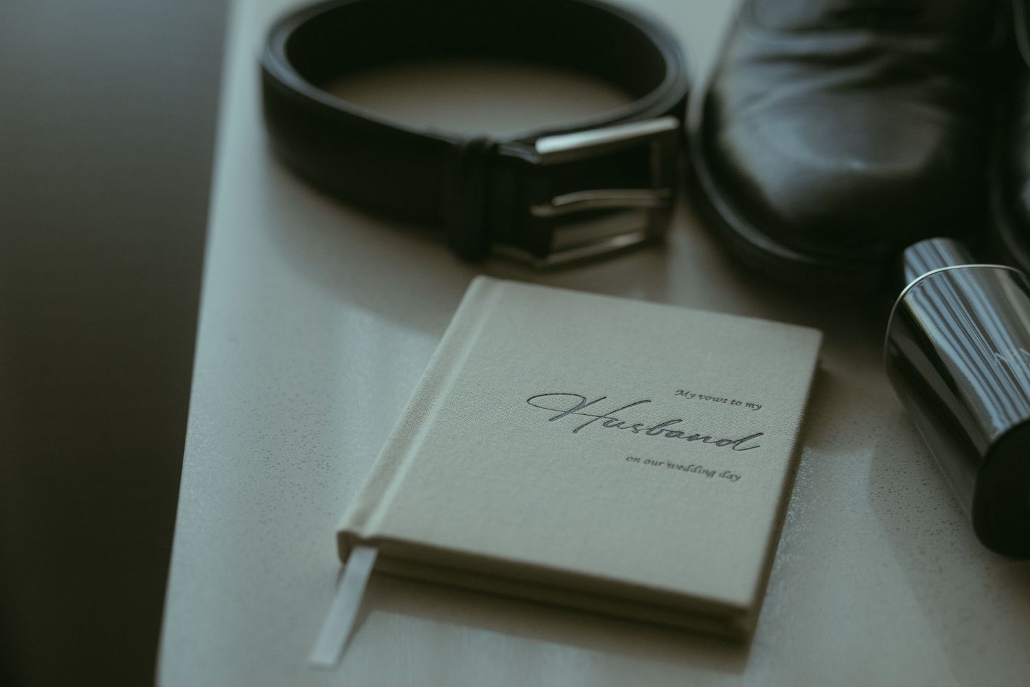 Close-up of groom wedding details including a leather belt, dress shoe, cologne, and a wedding vow booklet on a white surface.