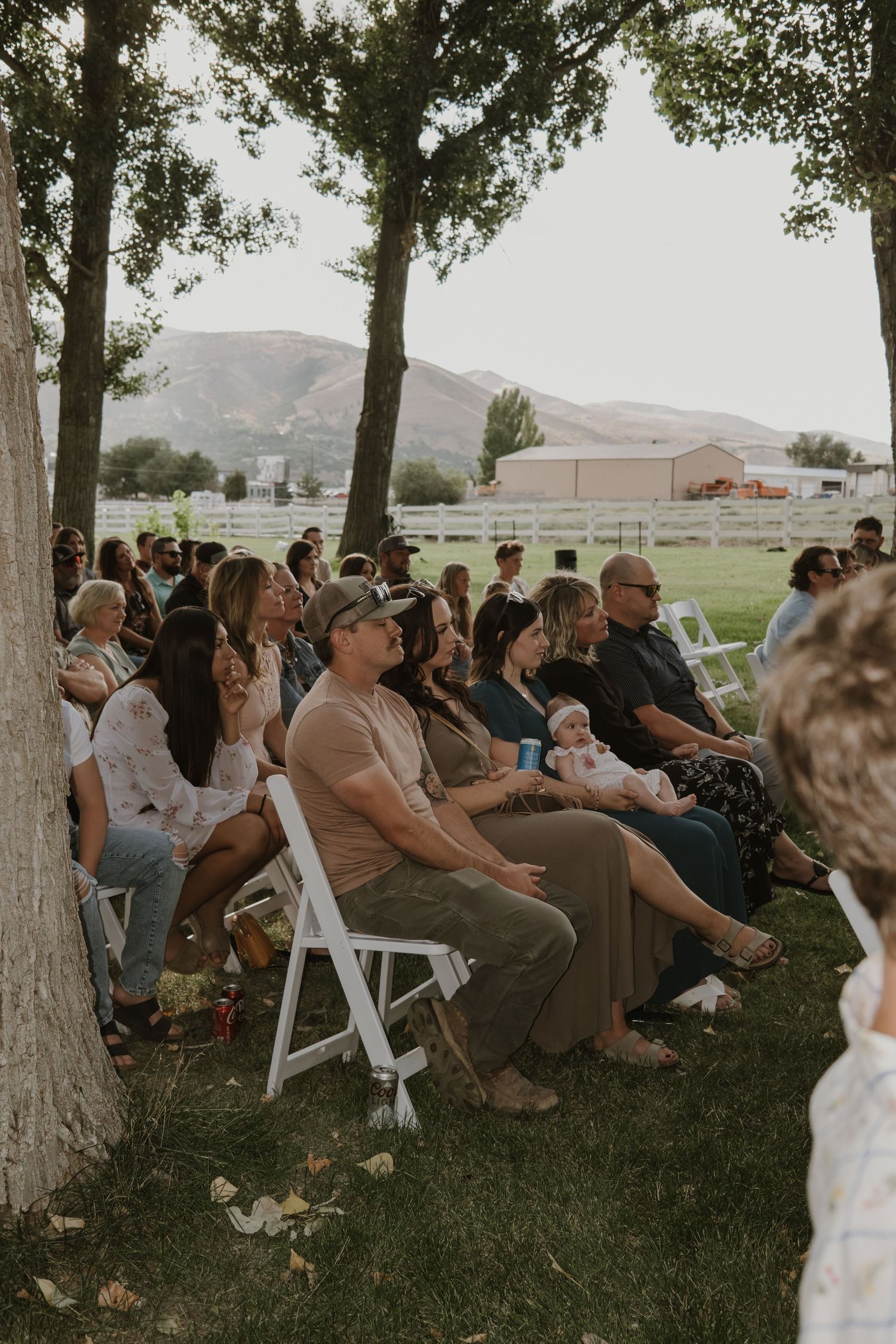 Wedding guests seated on white chairs under a canopy of trees at an outdoor ceremony venue.