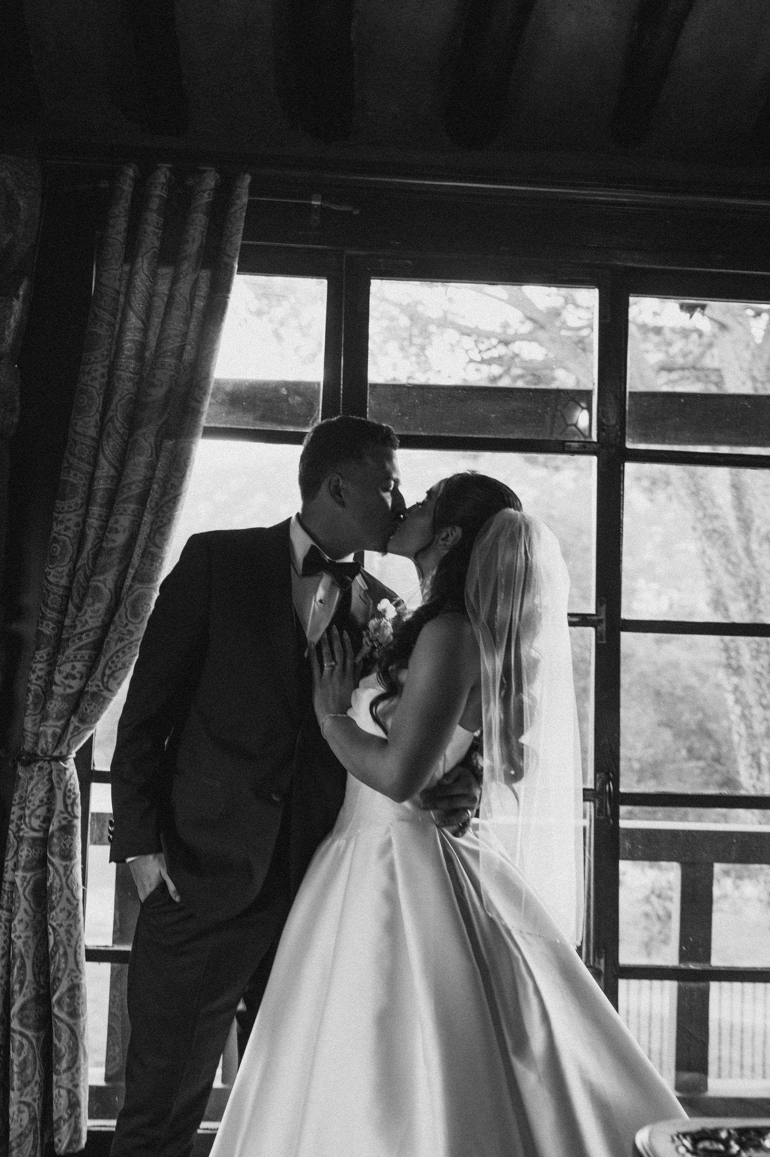 Black and white intimate moment between newlyweds by a window in formal attire.