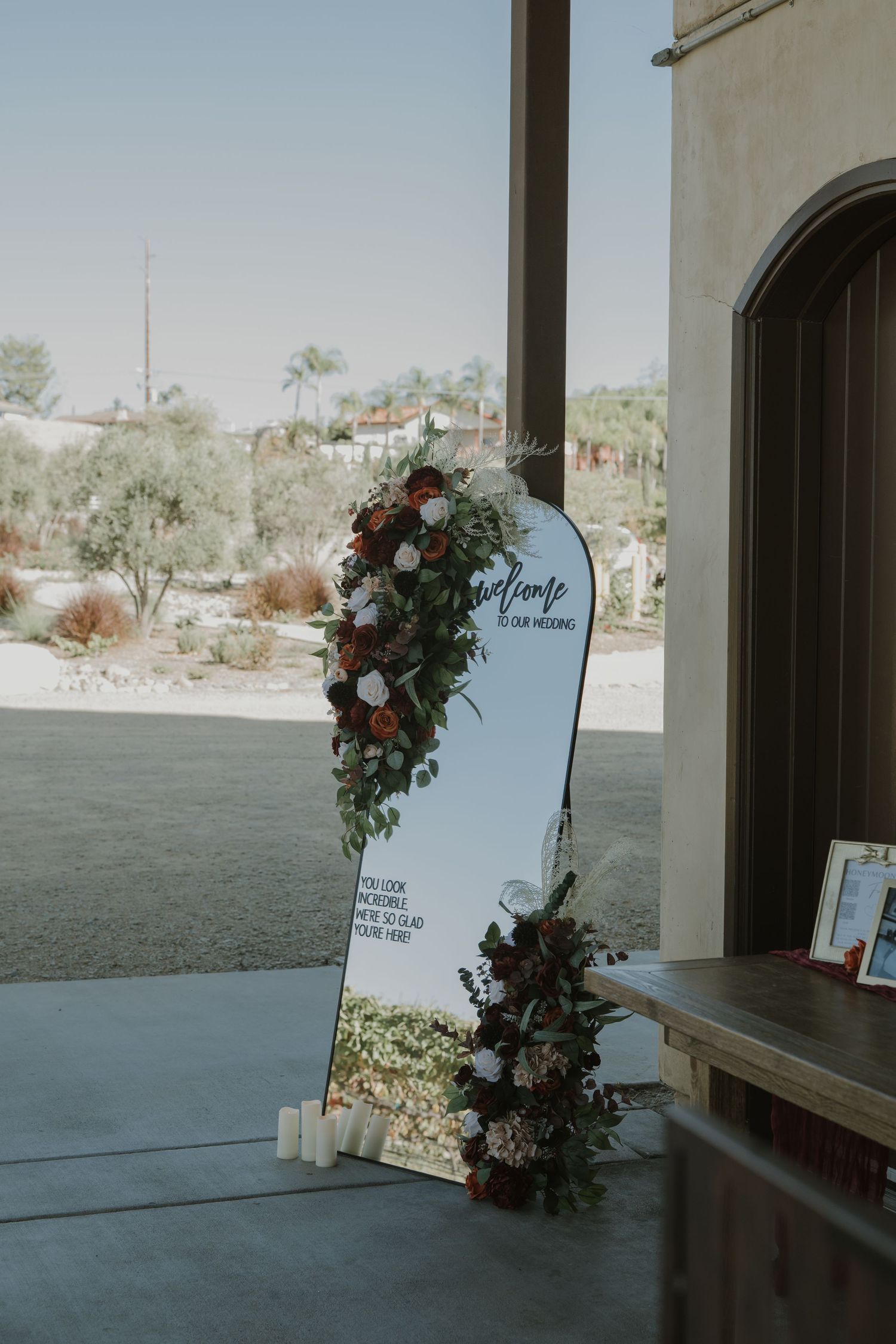 Wedding welcome sign decorated with floral arrangements against a white stucco wall.