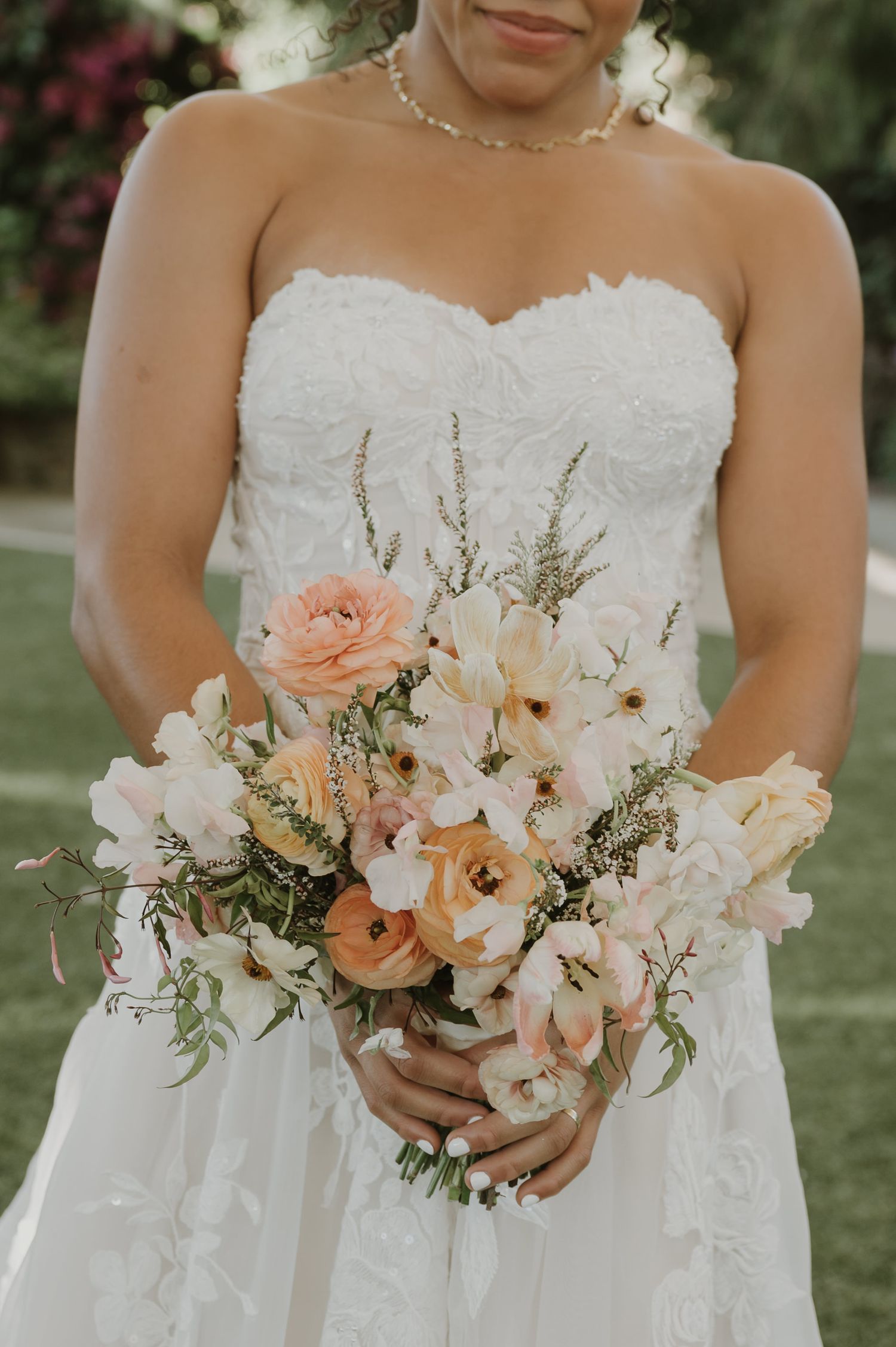 Bride in strapless lace wedding gown holding a romantic bouquet of peach ranunculus, white orchids, and blush blooms.