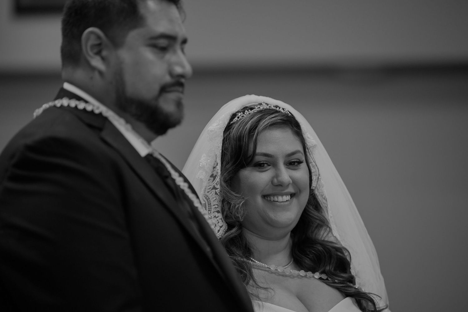 Black and white wedding photo of smiling bride in veil and tiara standing beside groom in suit during ceremony.