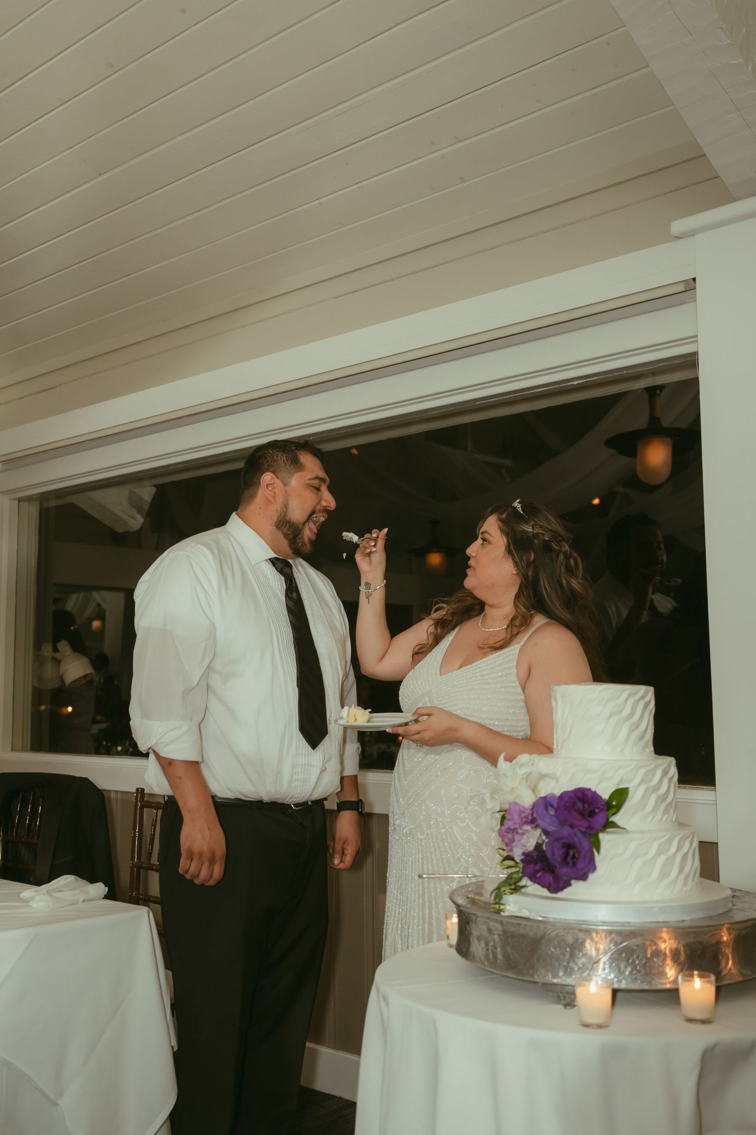 Bride feeding groom wedding cake slice at reception beside white tiered cake with purple flowers.