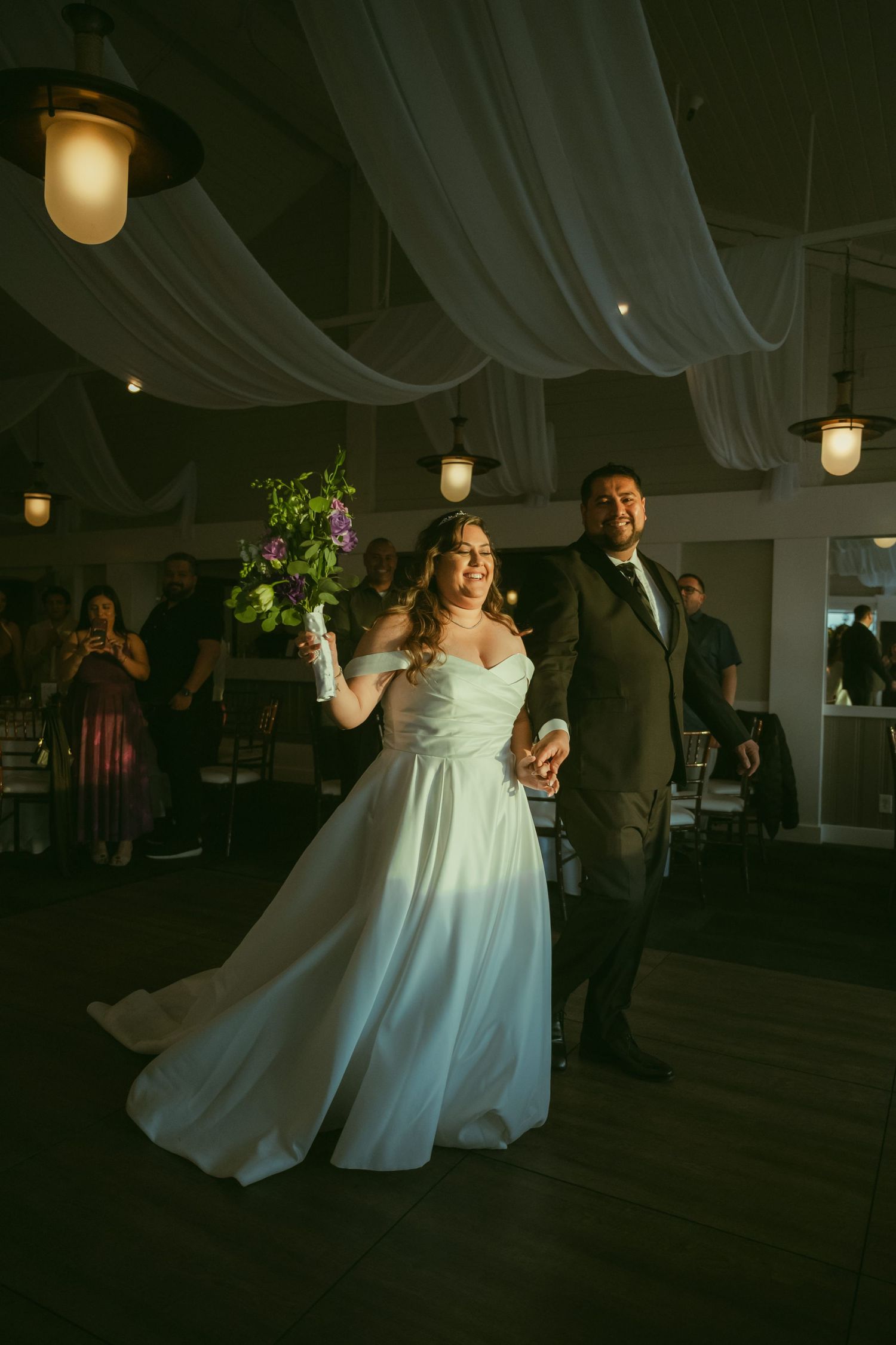 Bride and groom make grand entrance at wedding reception, bride holding purple bouquet in elegant white gown.
