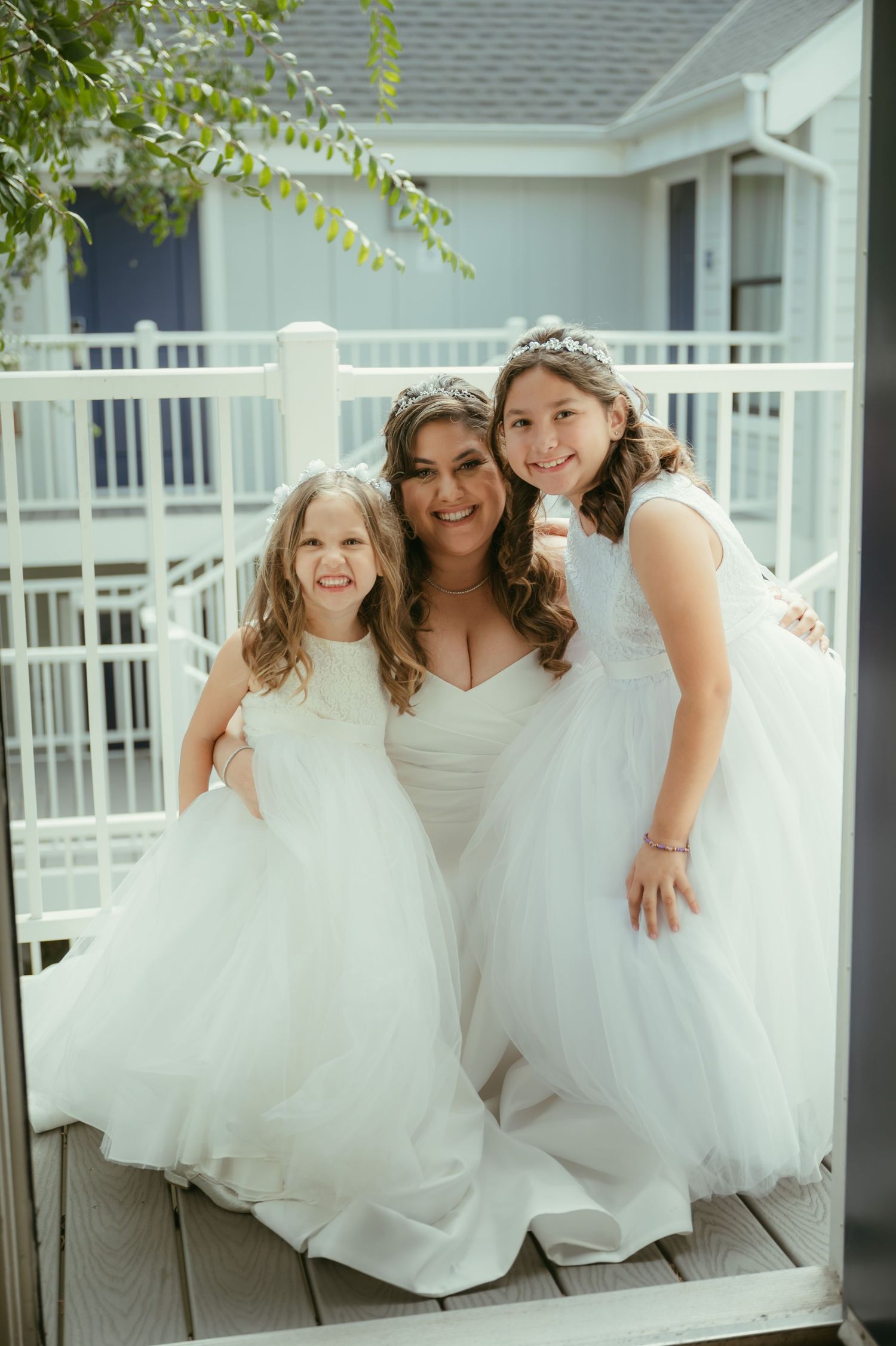 Bride in strapless wedding gown poses with two flower girls in white dresses on outdoor deck.