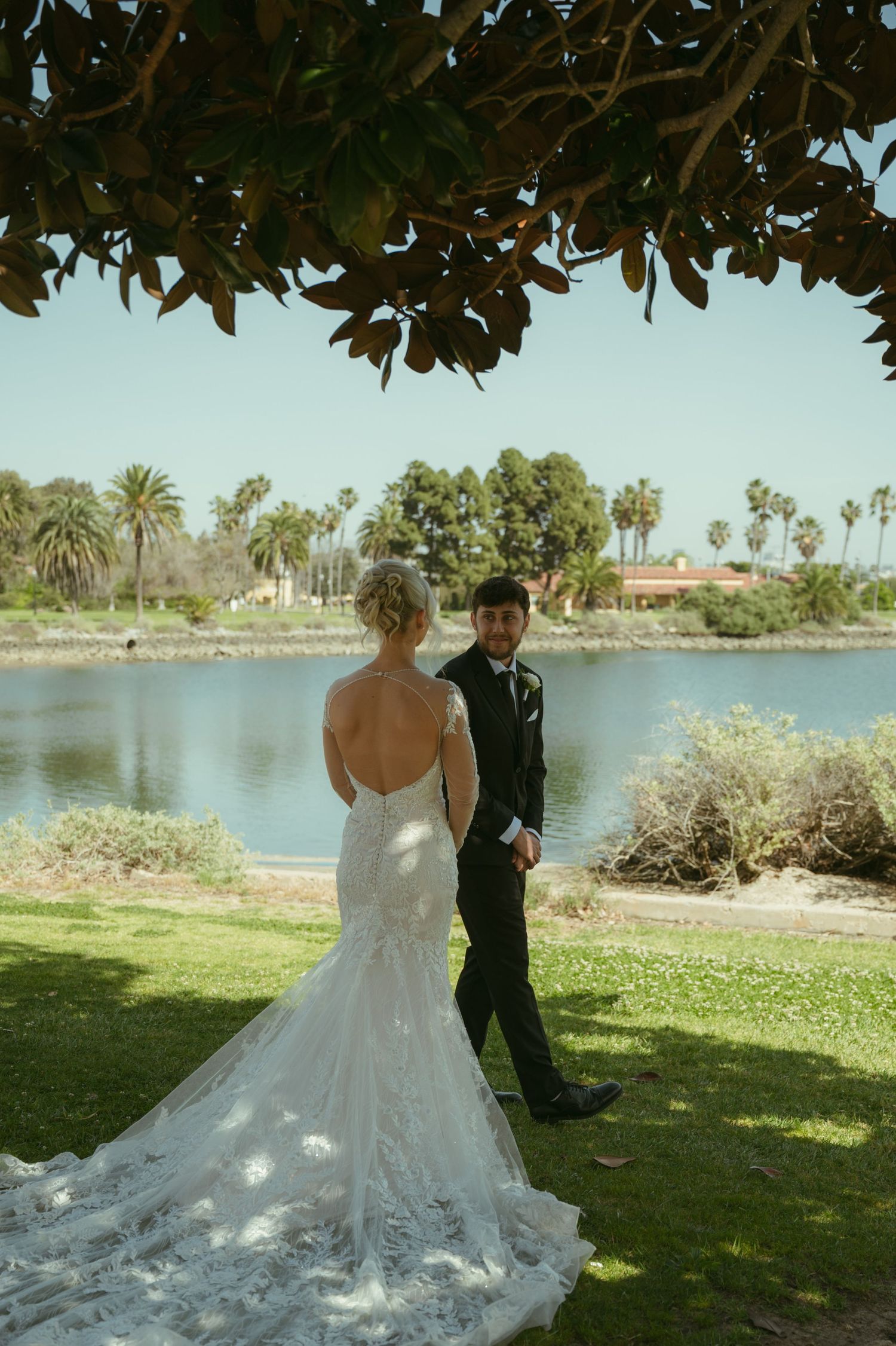 Bride in lace backless gown meets groom by a serene lakeside during first look.