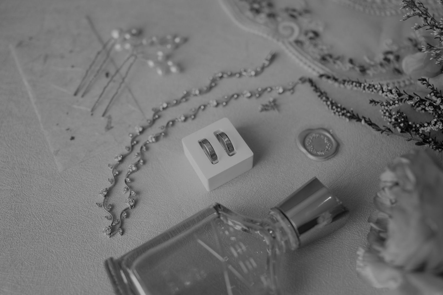 Black and white flat lay of perfume bottle, jewelry chain, hair pins, and small white box on white surface.
